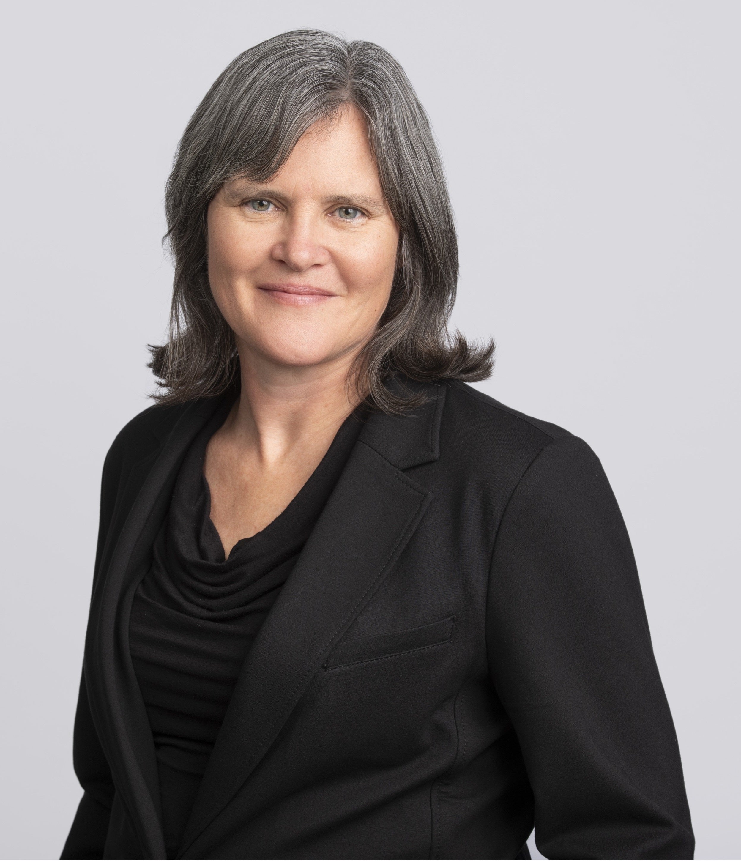 A profile photo of a smiling person with dark hair who is wearing a black shirt and blazer.
