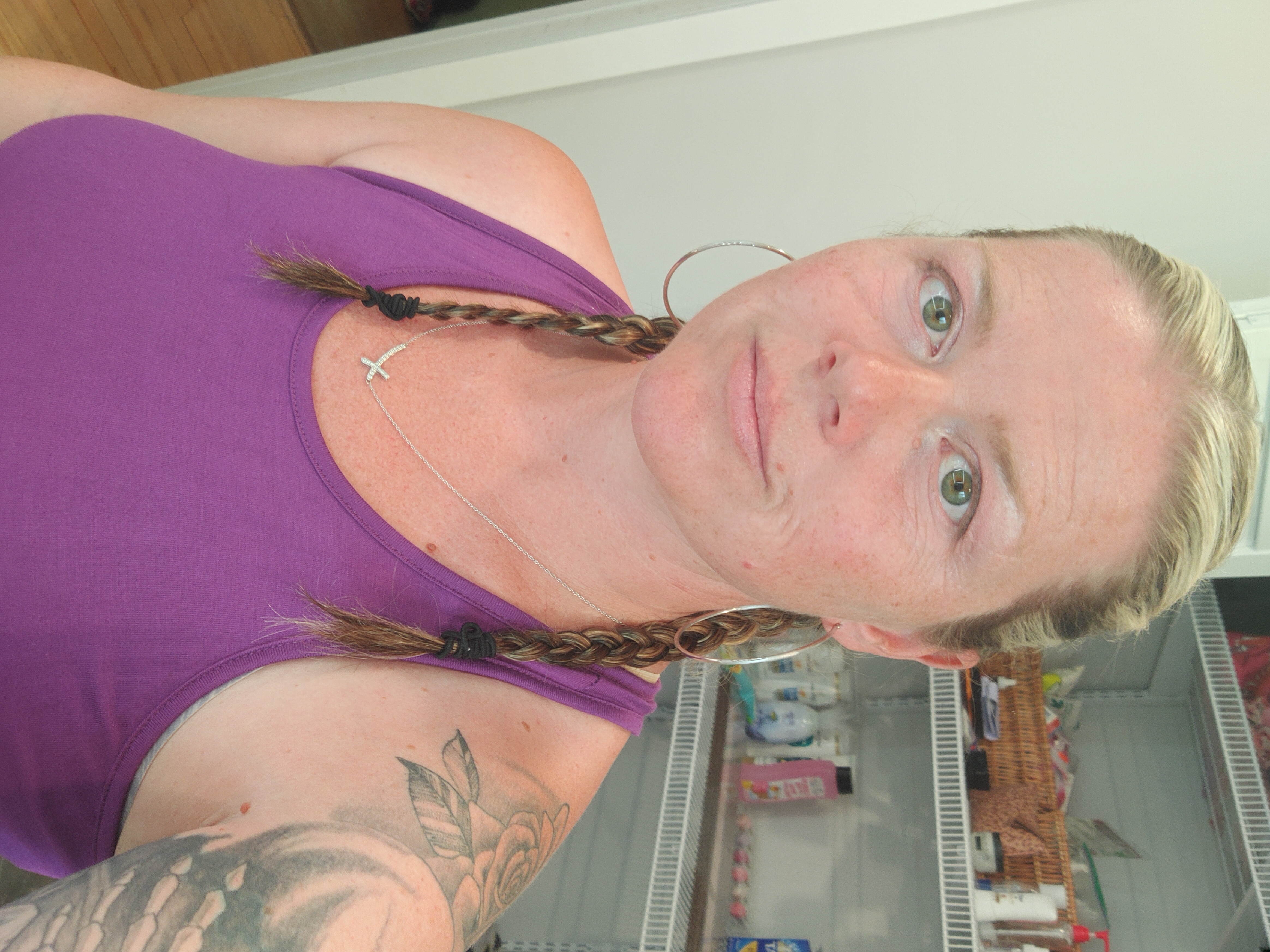 A person taking a selfie with blonde braids, a pink tank top, and tattoos on their left arm.
