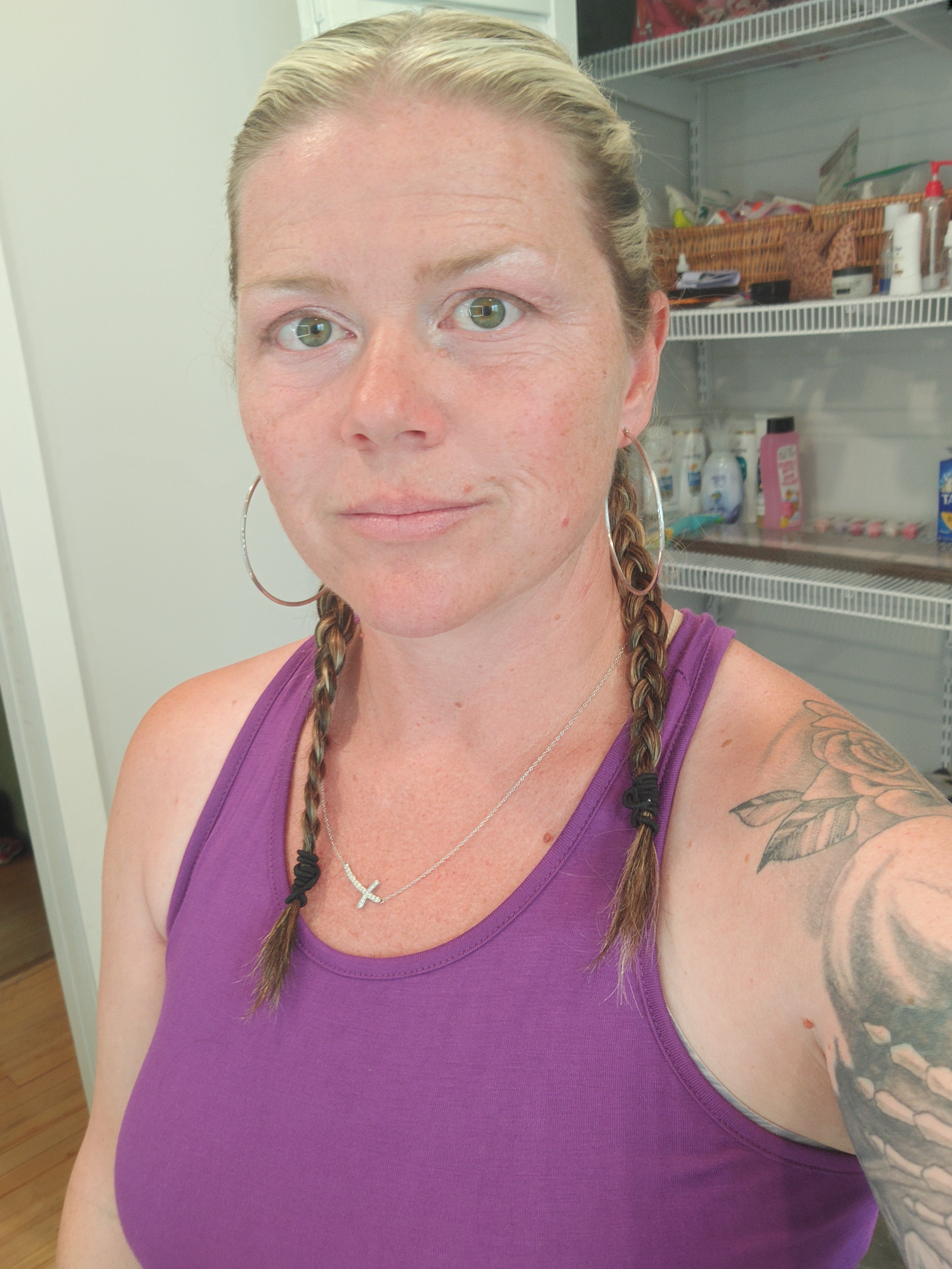 A person taking a selfie with blonde braids, a pink tank top, and tattoos on their left arm.