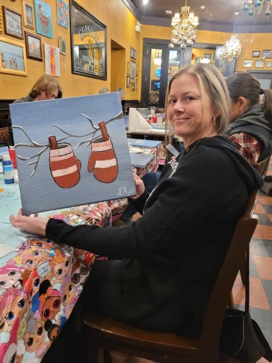 Woman smiles holding a painting of two mittens hanging on a branch. 