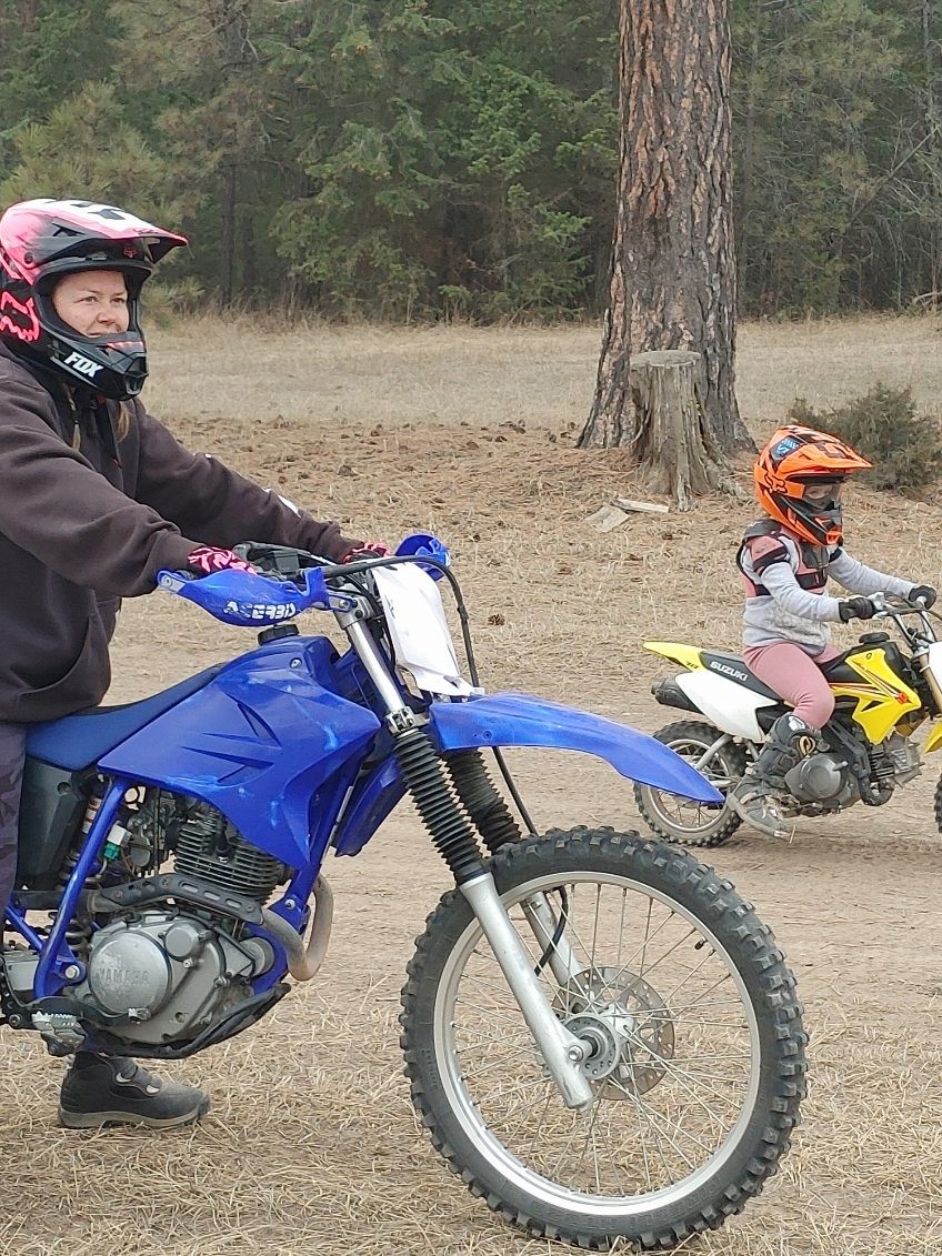 Woman wearing a black hoodie, pink helmet, and pink gloves sits on a blue dirtbike. Child in pink and grey outfit and orange helmet sits on a yellow and black dirtbike.