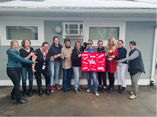 A group of 11 people wearing warm clothing stands in front of a blue grey building and point at a red poster held by one of the people