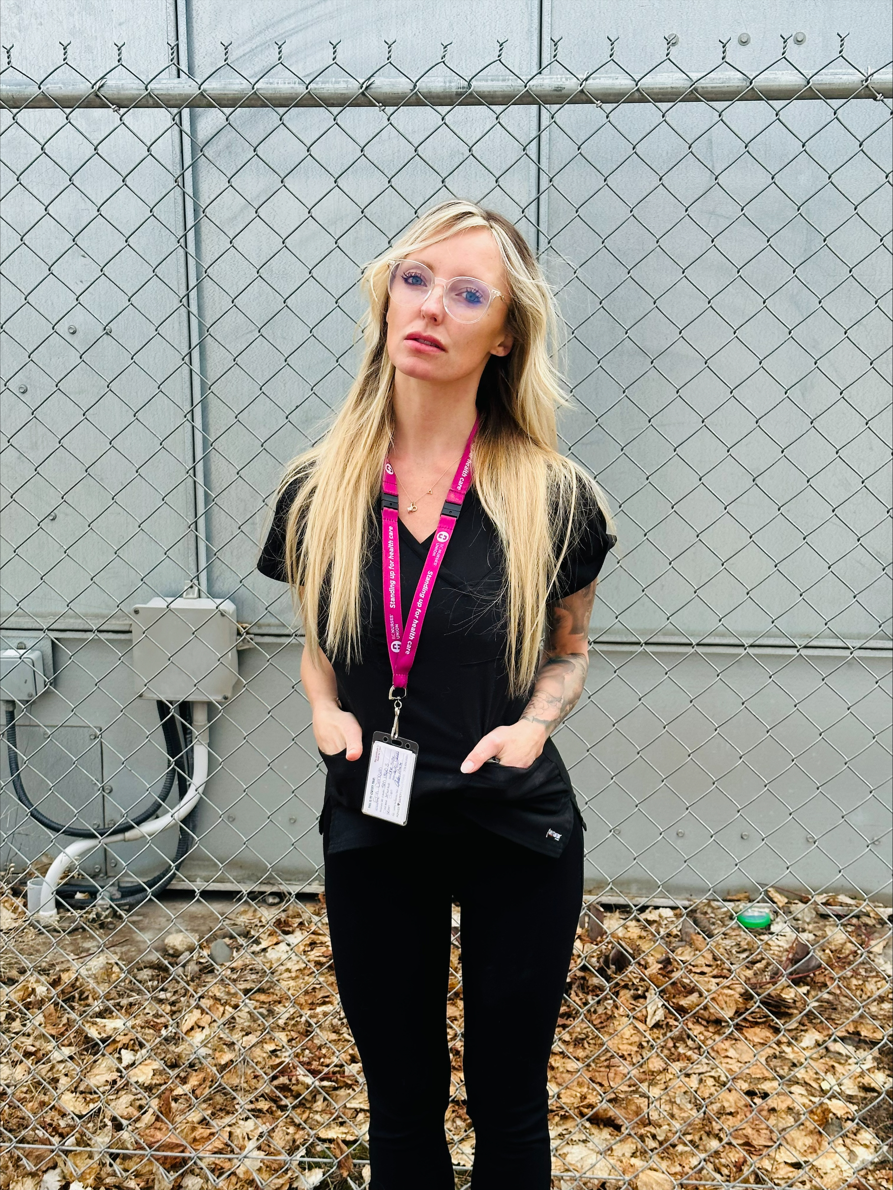 A person in black tshirt and pants with long blonde hair stands with their hands in their pockets in front of a chain link fence.