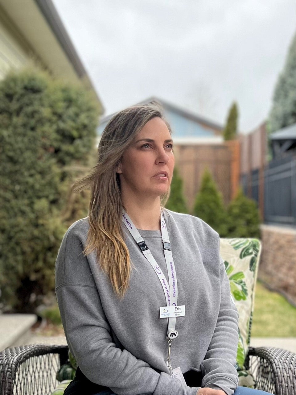 A person with long dark blonde hair in a grey sweatshirt and blue jeans sits on a garden chair in front of a hedge and fence.