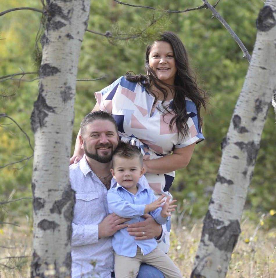 A woman with long brown hair in a white dress stands behind a man in a white dress shirt sitting between two birch trees and holding a young boy.