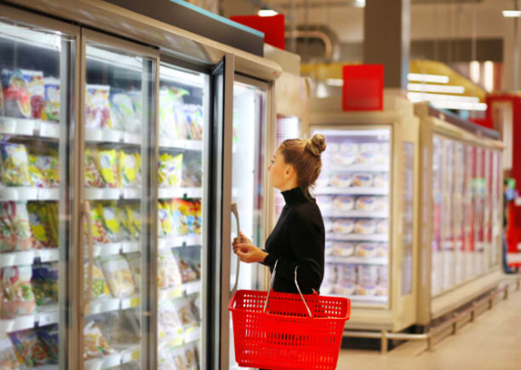 A girl with a hair bun and wearing a black turtleneck holds a red grocery basket in front of a freezer at a grocery store