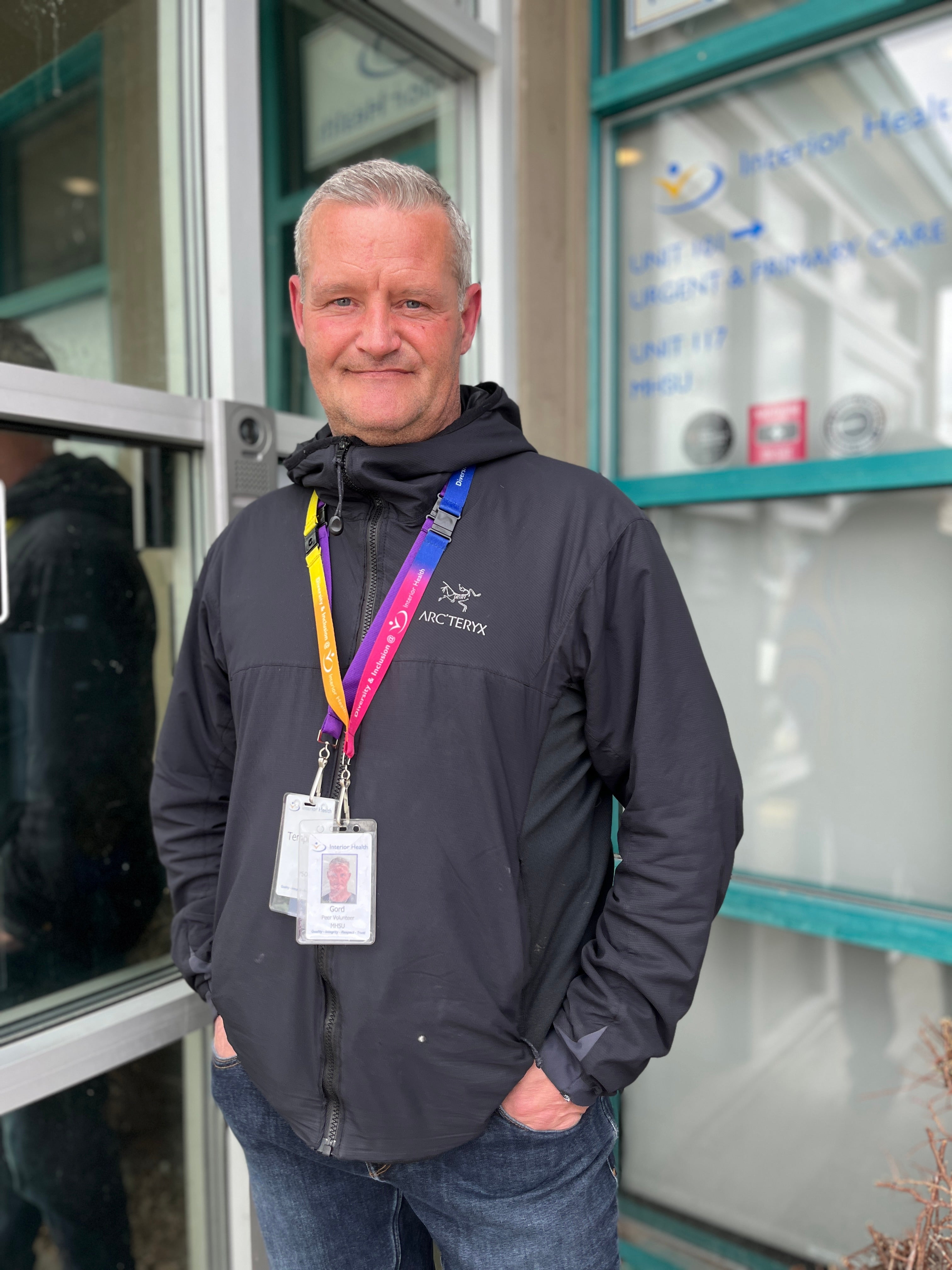 A person stands in a black jacket with a multi-coloured lanyard and tags around their neck in front of a building window.