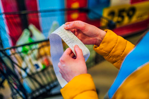 Two hands of a person wearing a yellow puffy coat holding a grocery bill in front of a grocery cart