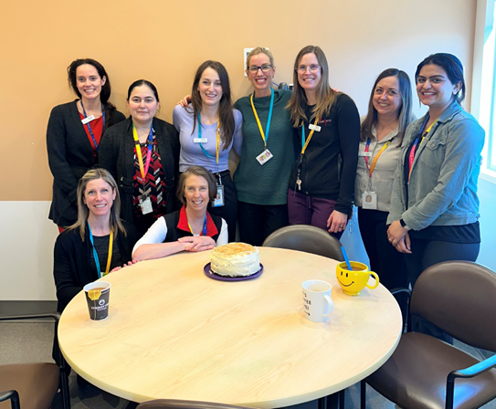 A group of 9 women with lanyards and wearing business casual clothes stand around a table with a cake on it