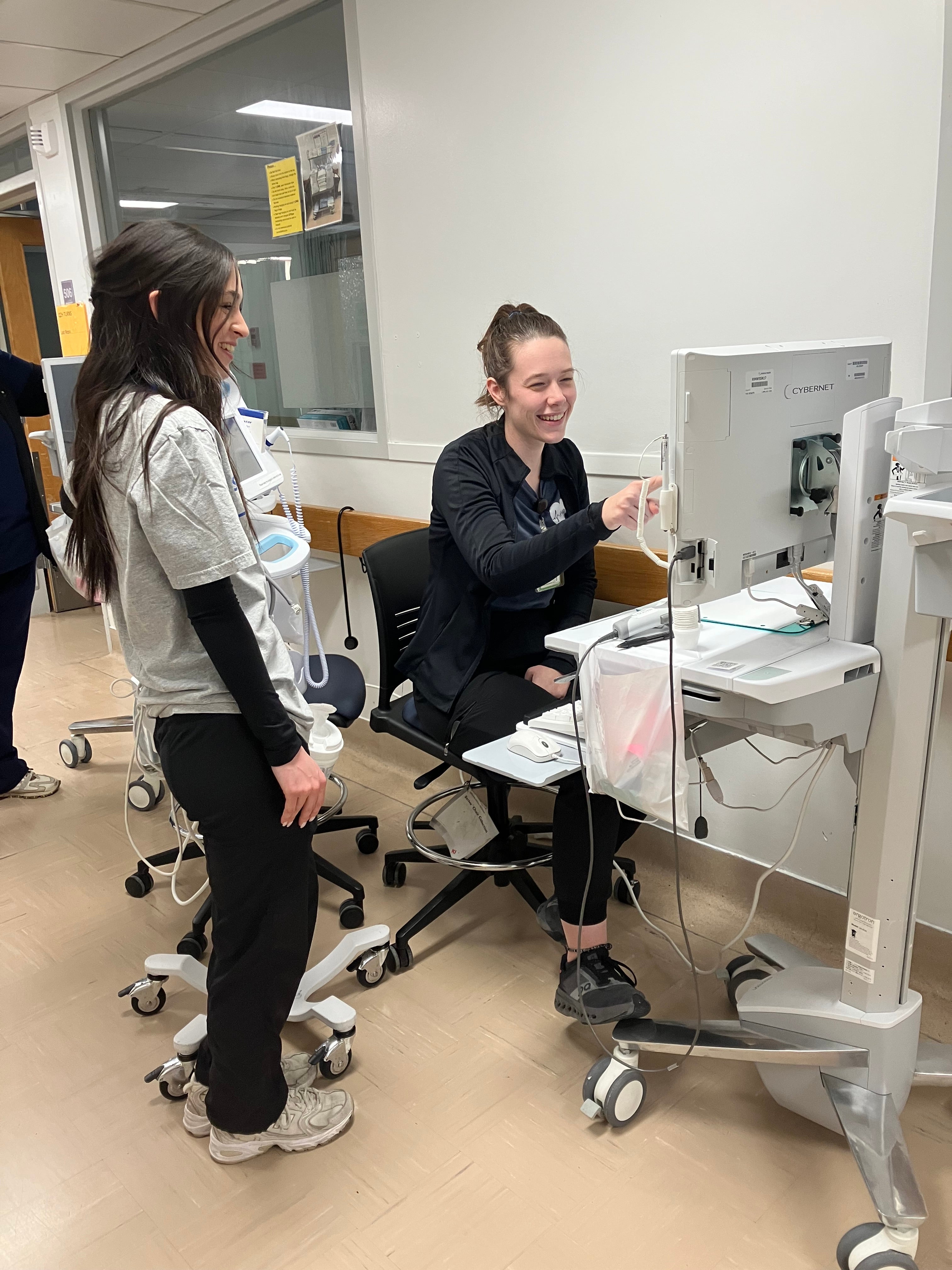 A person sitting in a chair points at a monitor while a young person with long brown hair looks on. They are in a hospital hallway.
