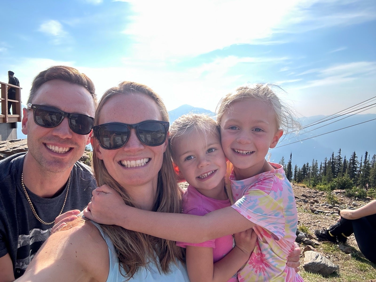 A smiling family of four including a mom and dad in sunglasses and two toddlers takes a selfie on a sunny day