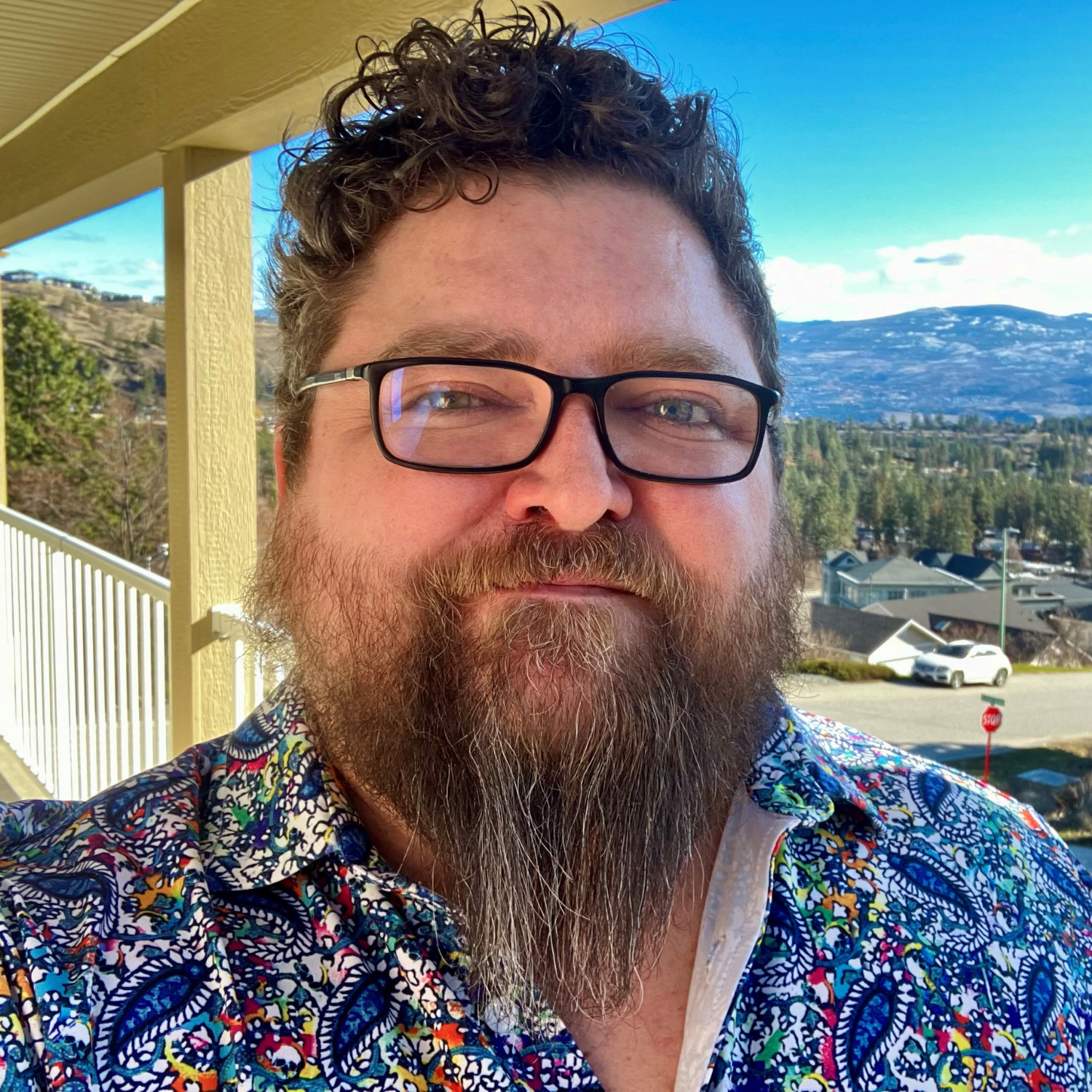 a person with glasses and a blue shirt and brown hair and a beard looks to camera