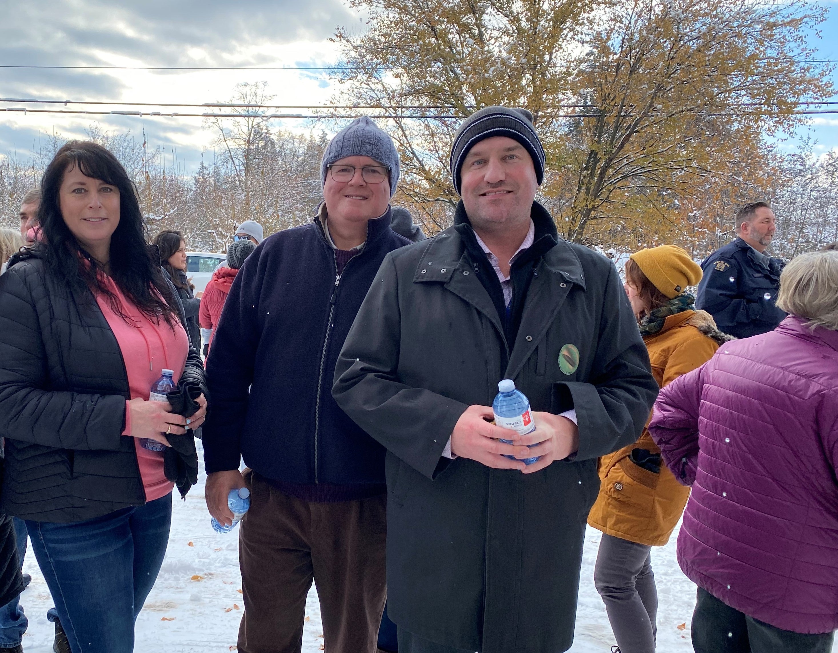 Three people wearing dark winter coats and hats smiling at an outdoor event.