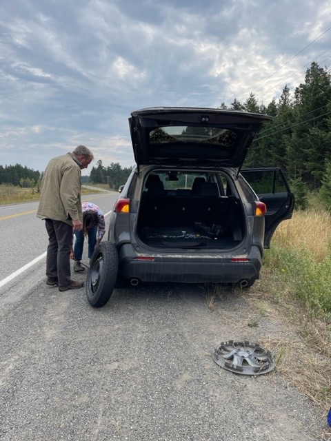 A vehicle pulled over on the shoulder of the road gets its tire replaced by one person while another person watches.