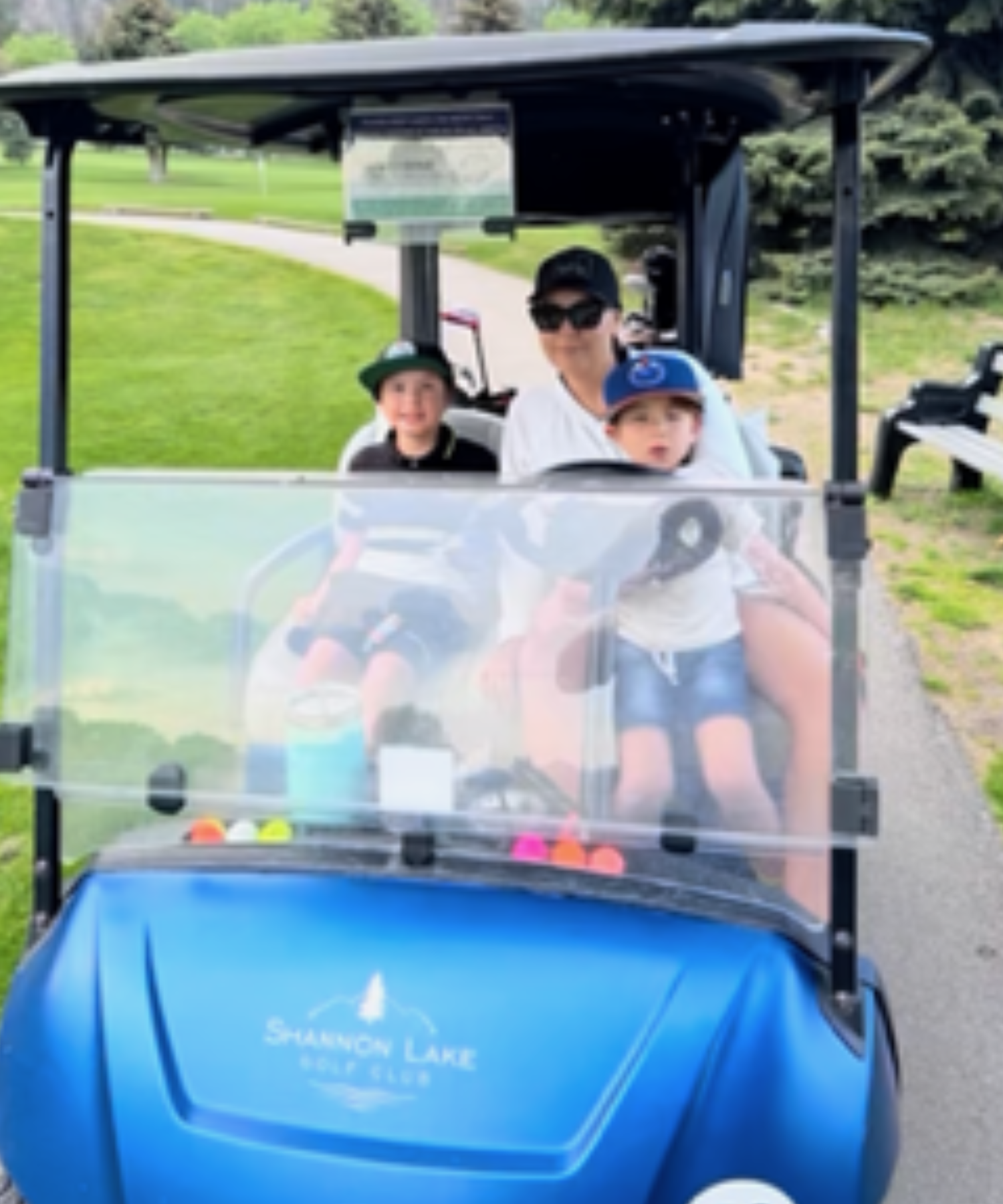 A woman and two young boys drive a blue golf cart through a golf course with trees and mountains in the background. 