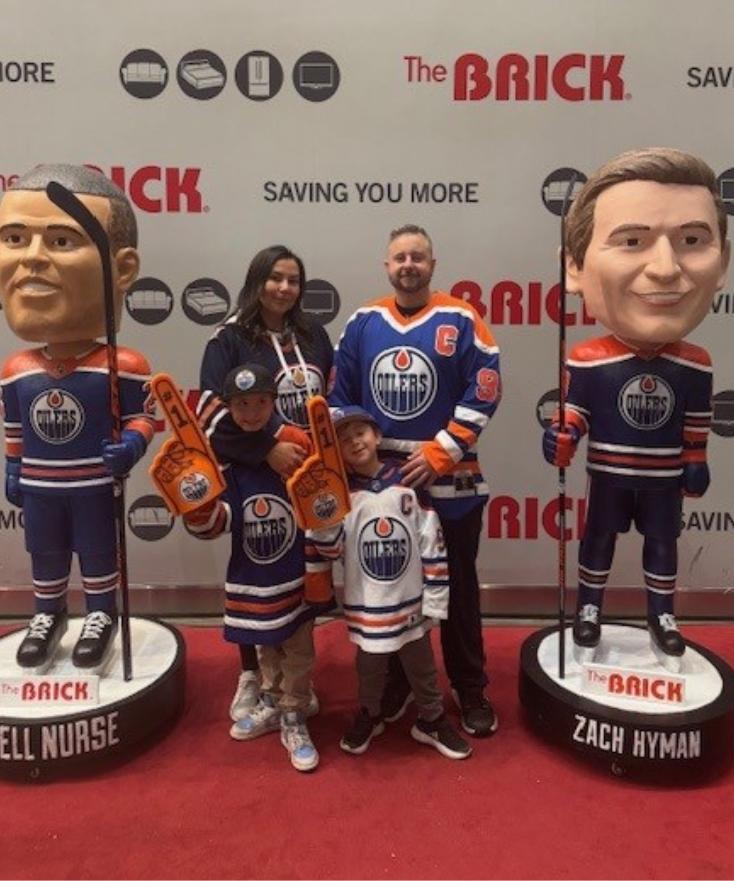 A woman, man, and two children wearing Edmonton Oilers jerseys stand between two life-size Edmonton Oilers bobble heads. The children hold up foam fingers. 
