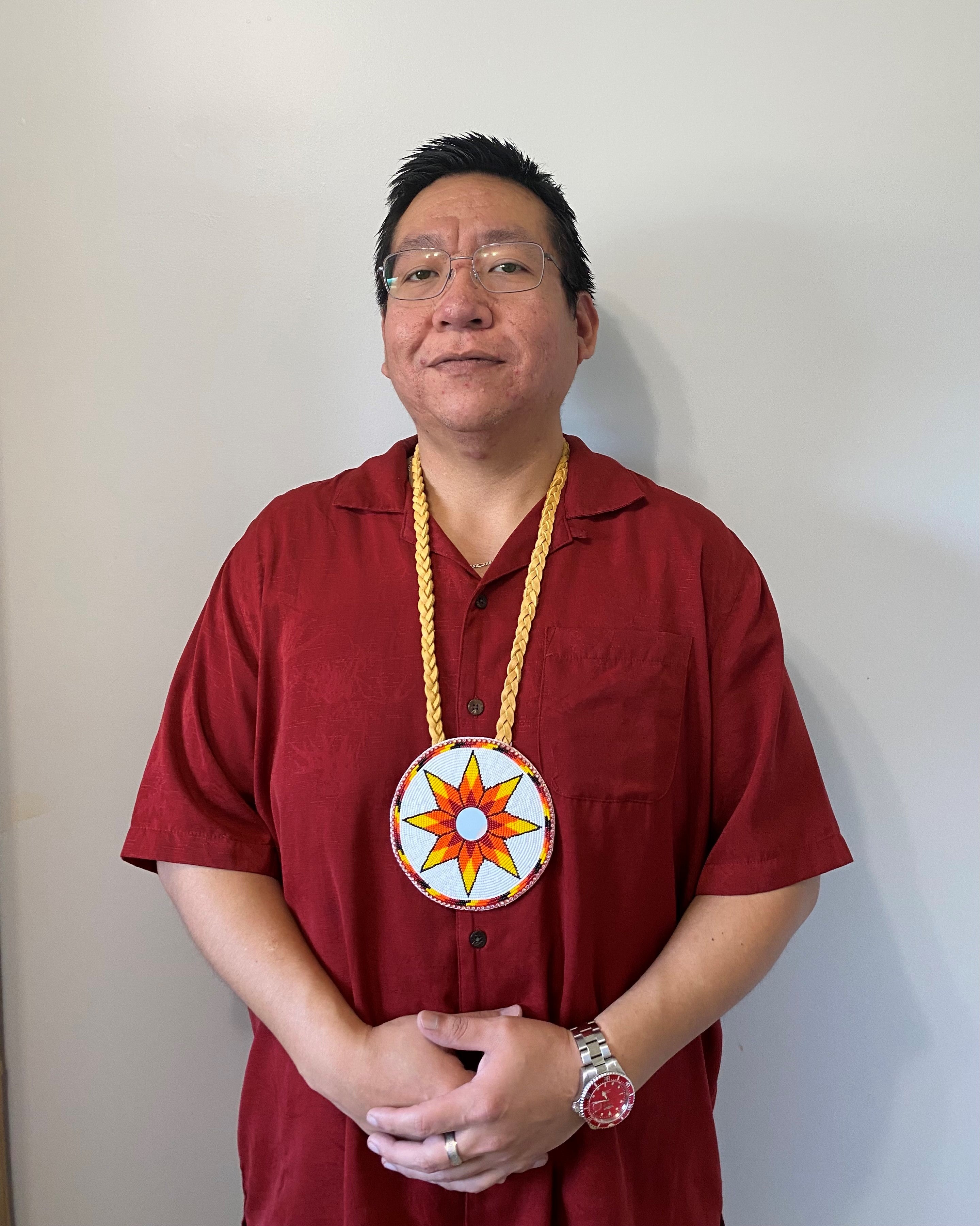 A person with dark wavy hair and glasses wearing a red button up shirt and long necklace that holds a circular pendant with an orangey-yellow flower petal.
