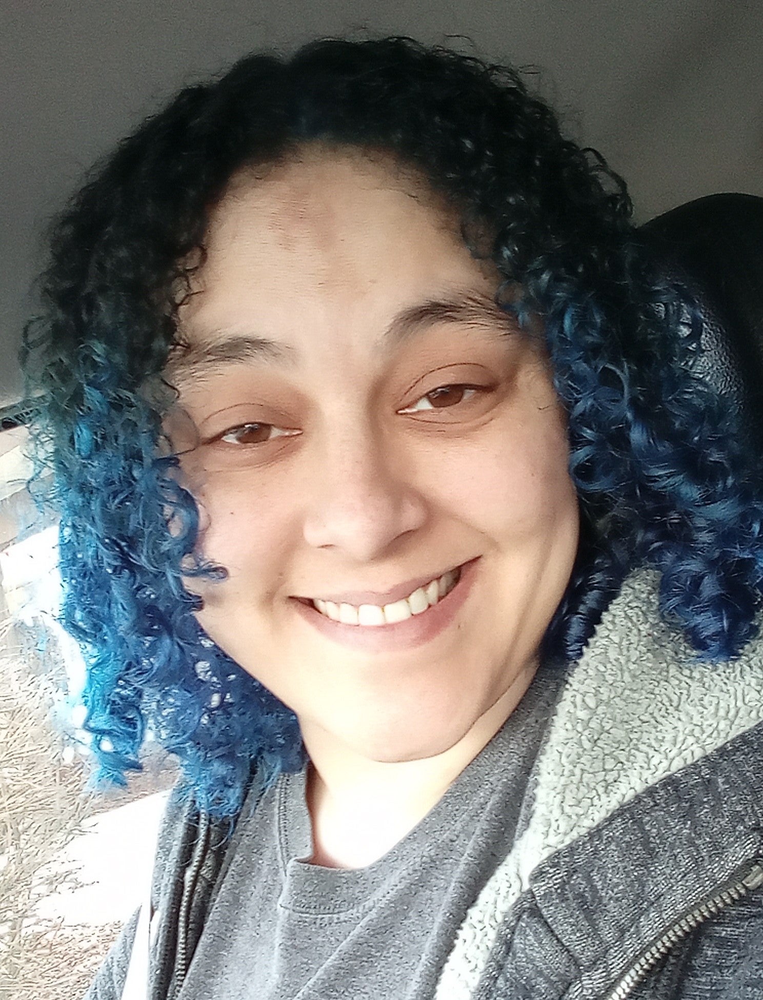 A person with medium length curly hair dyed blue wearing a grey hoodie and tshirt.