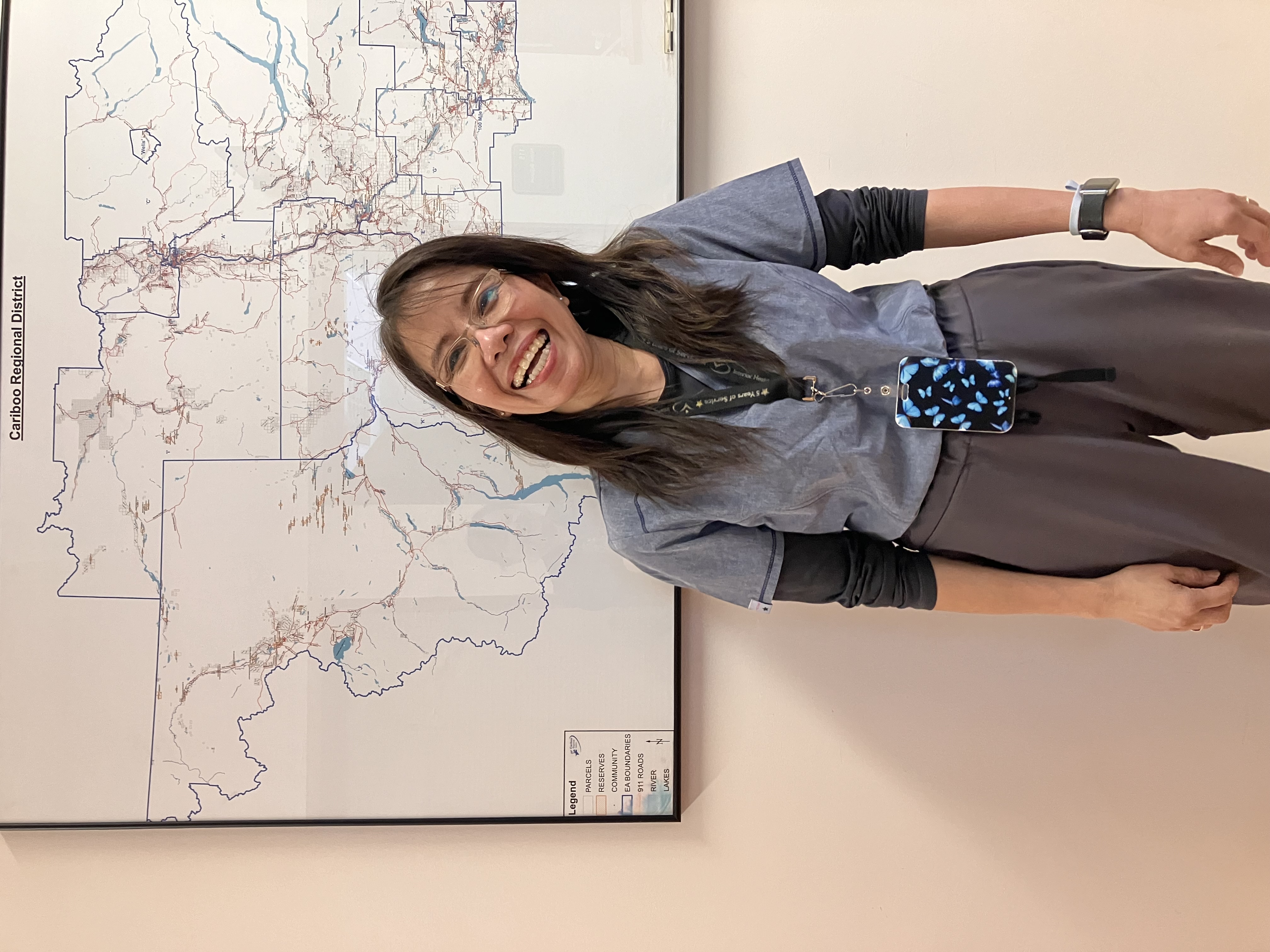 A smiling woman with long dark hair and glasses and grey medical clothes smiles in front of a map in an office
