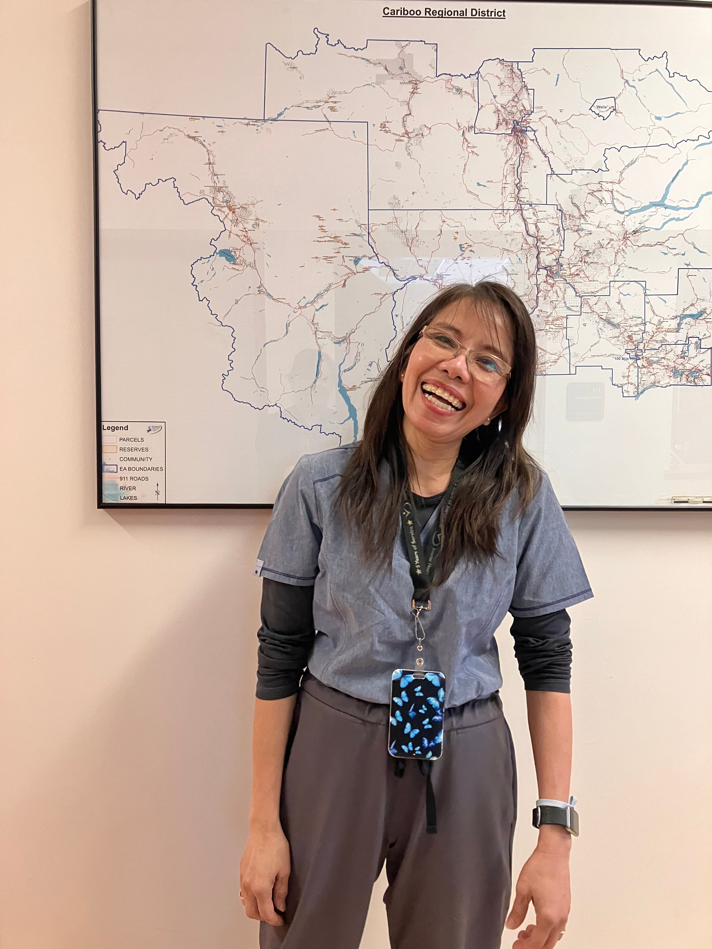 A smiling woman with long dark hair and glasses and grey medical clothes smiles in front of a map in an office