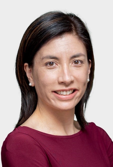 A smiling person with long dark hair tucked behind their ears, wearing small earrings and a burgundy top.