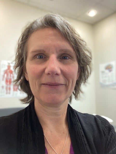 A smiling person with short grey hair wearing a black blouse in an office setting.