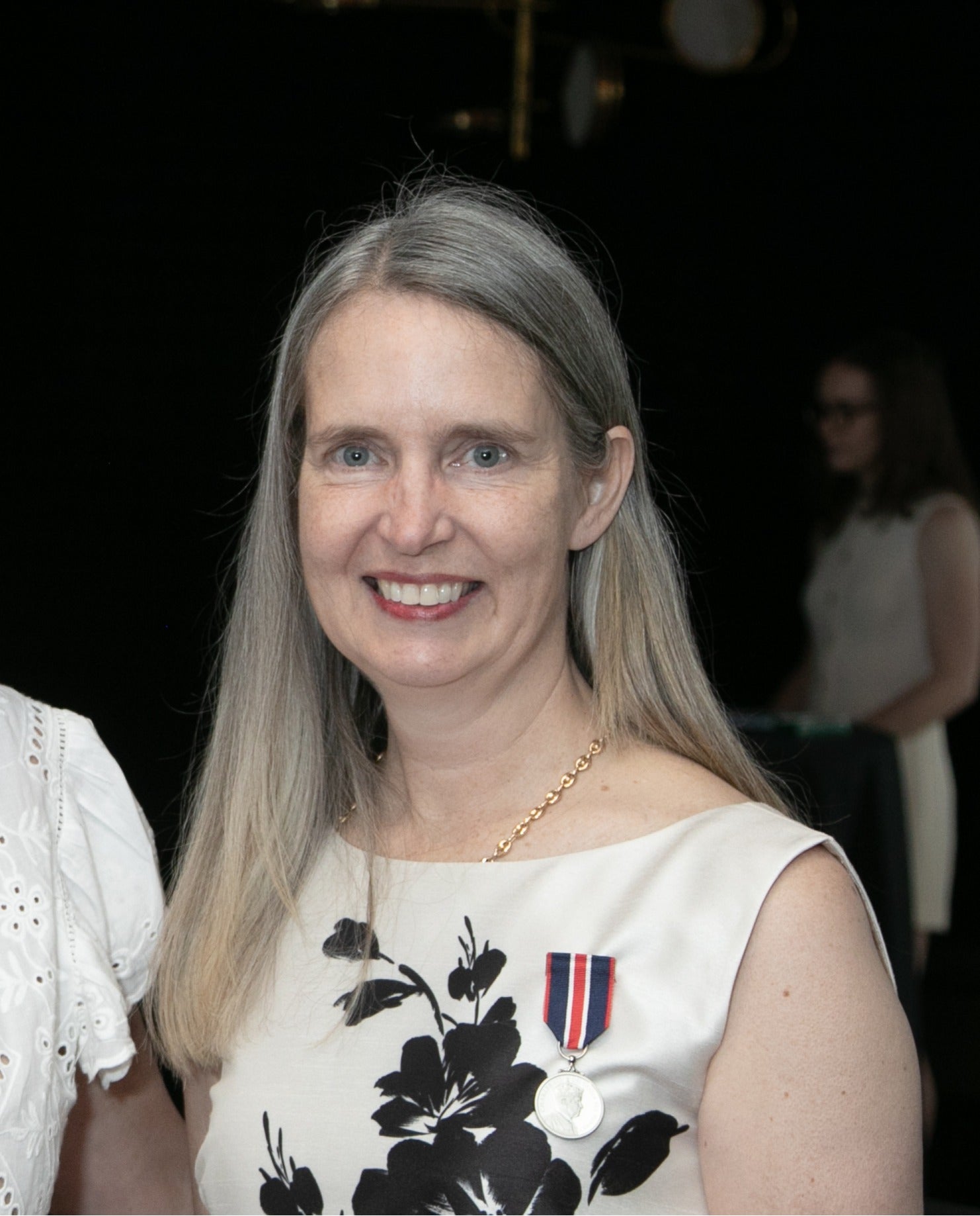 A smiling person with long grey-blonde hair in a white floral dress. On the dress is pinned a ribbon and medal.