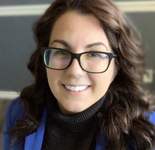 A woman with dark wavy hair and square glasses wearing a black shirt and blue scarf