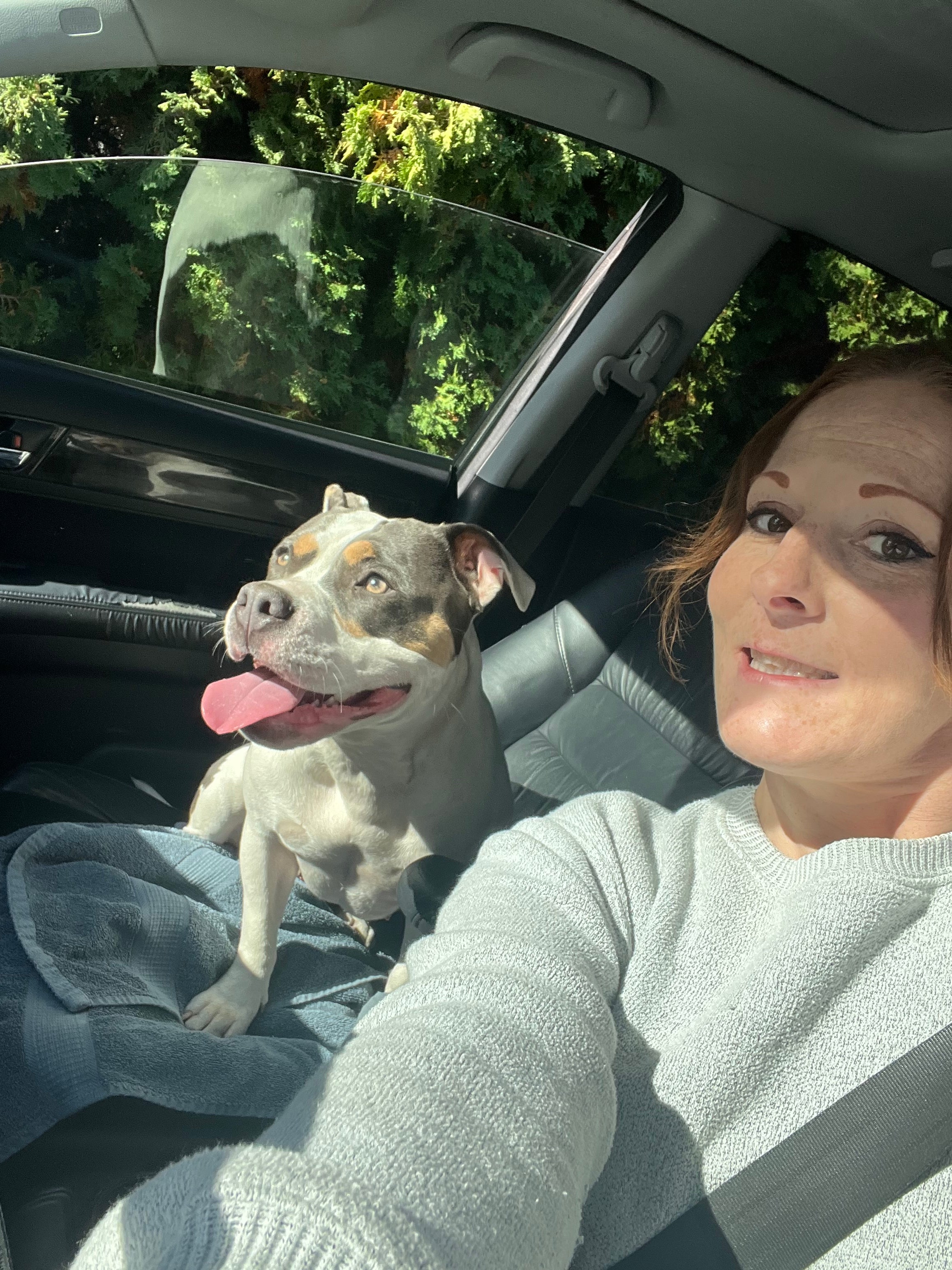 A selfie of a person in the driver's seat of a vehicle with a dog sticking its tongue out in the passenger seat.