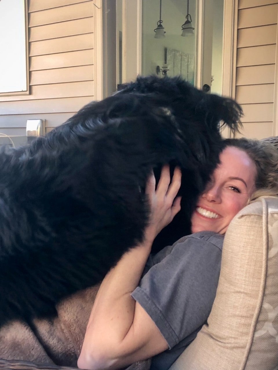 Person smiling joyfully while embracing a large black dog on a porch sofa.