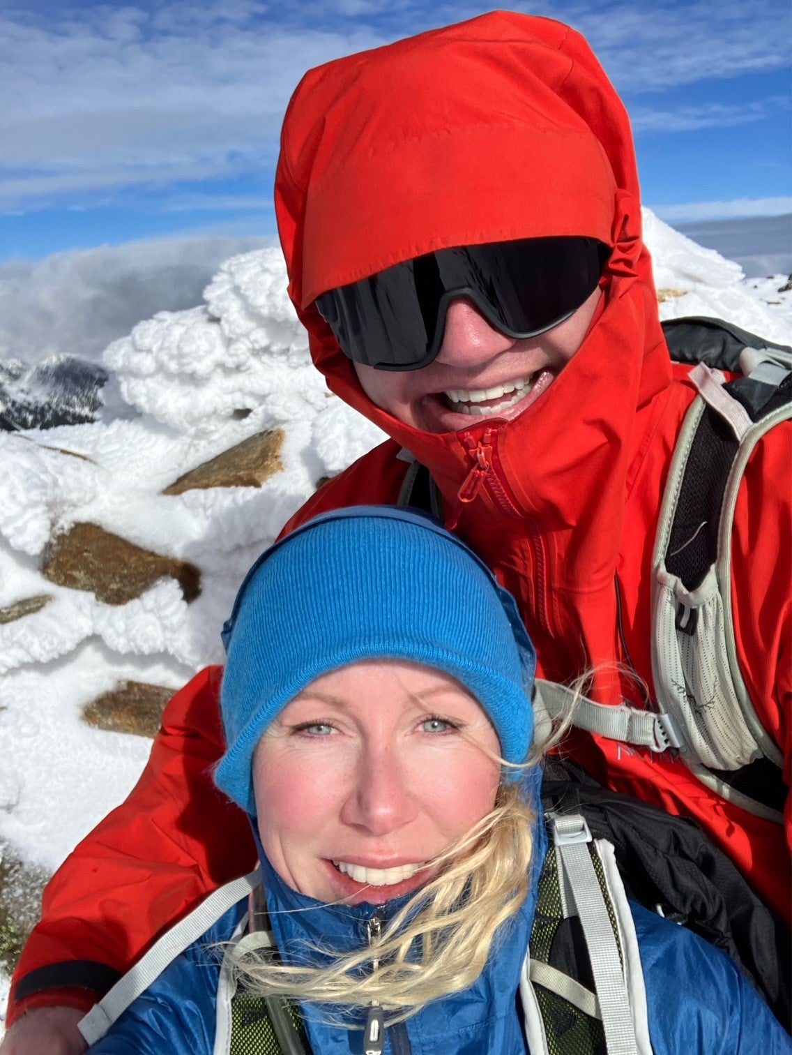 Two hikers in winter gear smiling for a selfie on a snowy mountain summit.