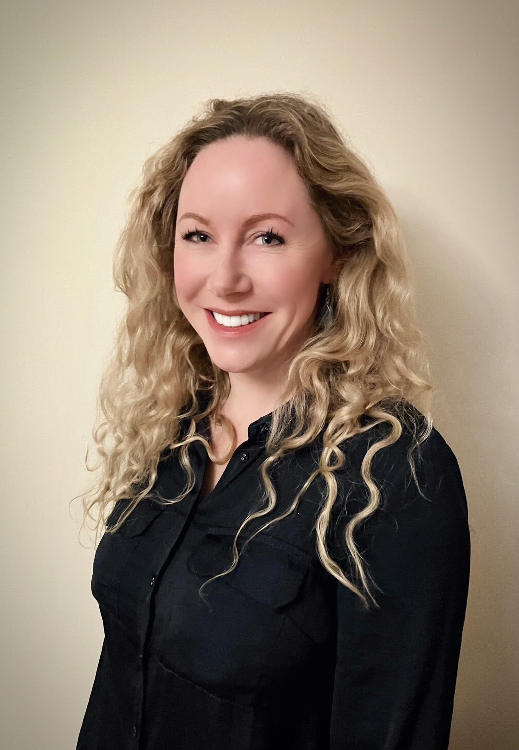 A smiling person with long curly blonde hair wearing a black shirt against a light background.