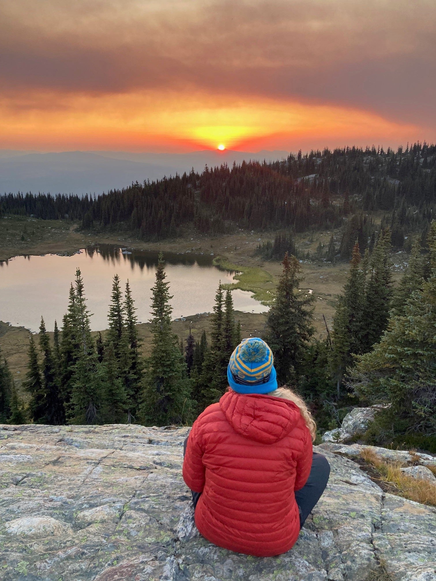 Person in a red jacket and blue hat sitting on a rock overlooking a scenic lake at sunset, with a forested mountain landscape in the background.