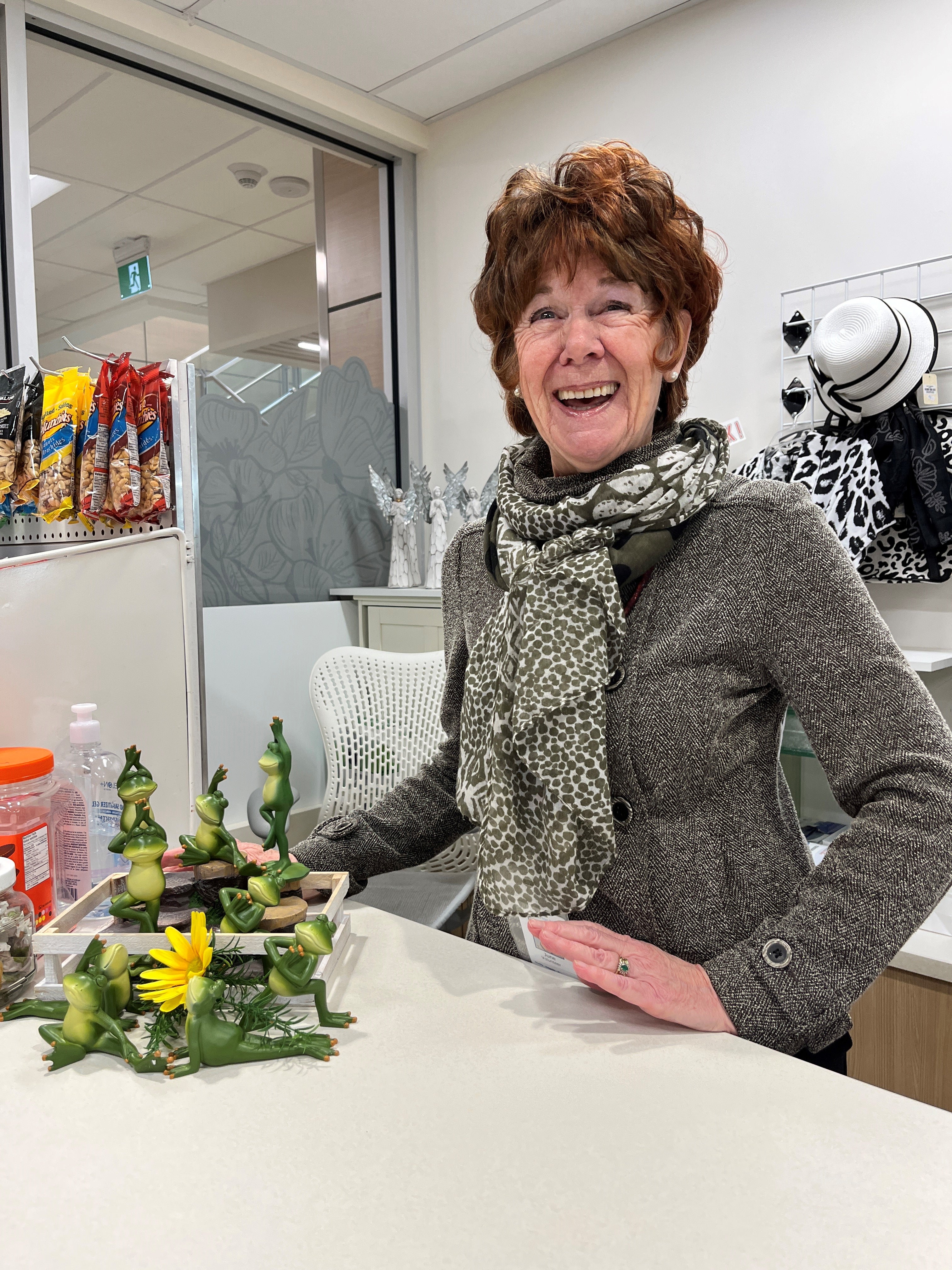 A person laughing and wearing a grey herringbone jacket and scarf stands behind a counter with a display of glass frog ornaments