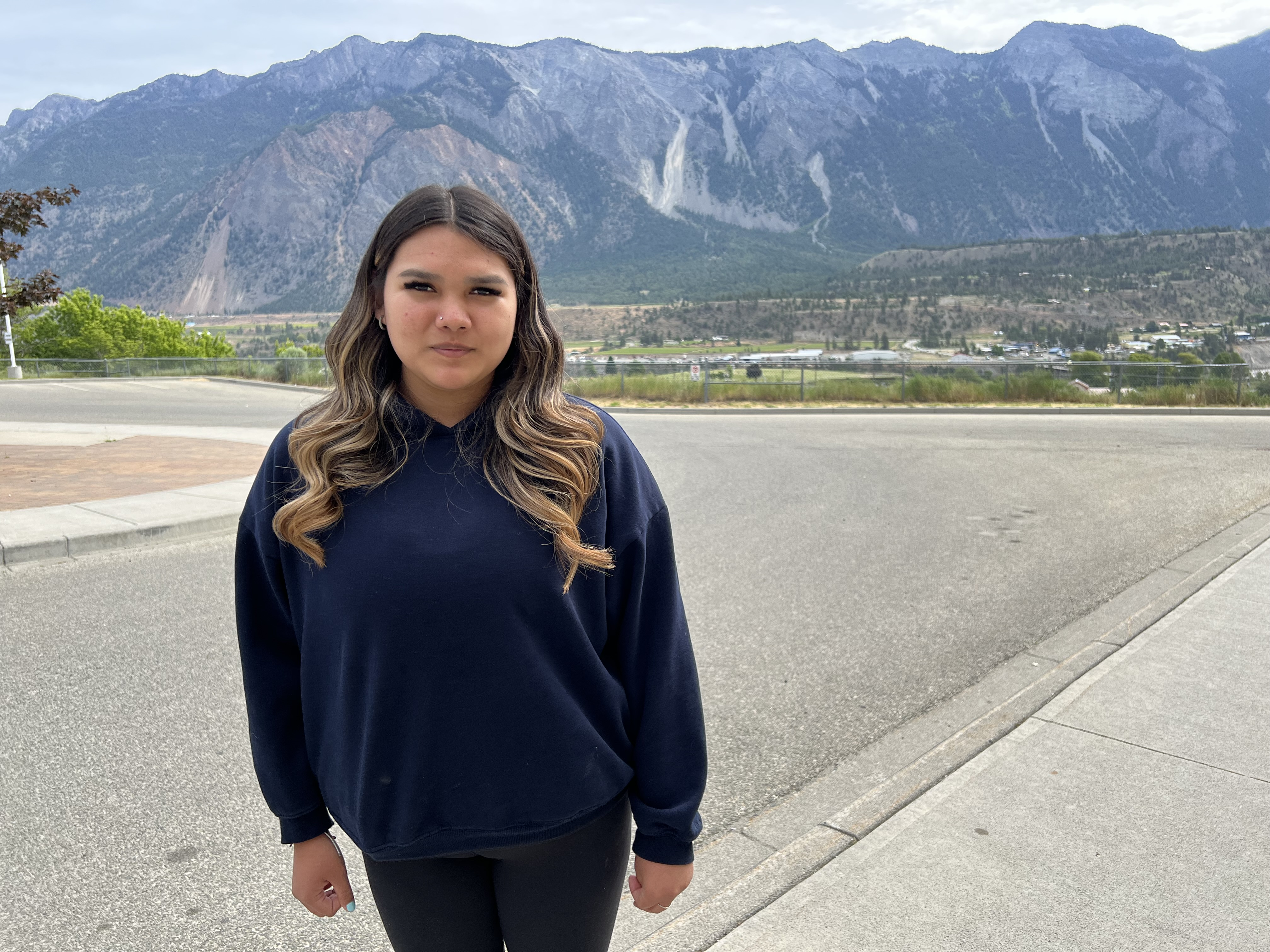 A girl with long brown blonde hair in a dark sweatshirt and pants stands in a parking lot with majestic mountains behind her.