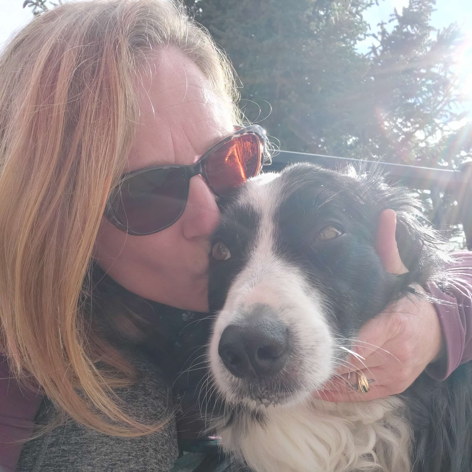 A blonde woman wearing sunglasses kisses her black and white dog in a selfie-style picture