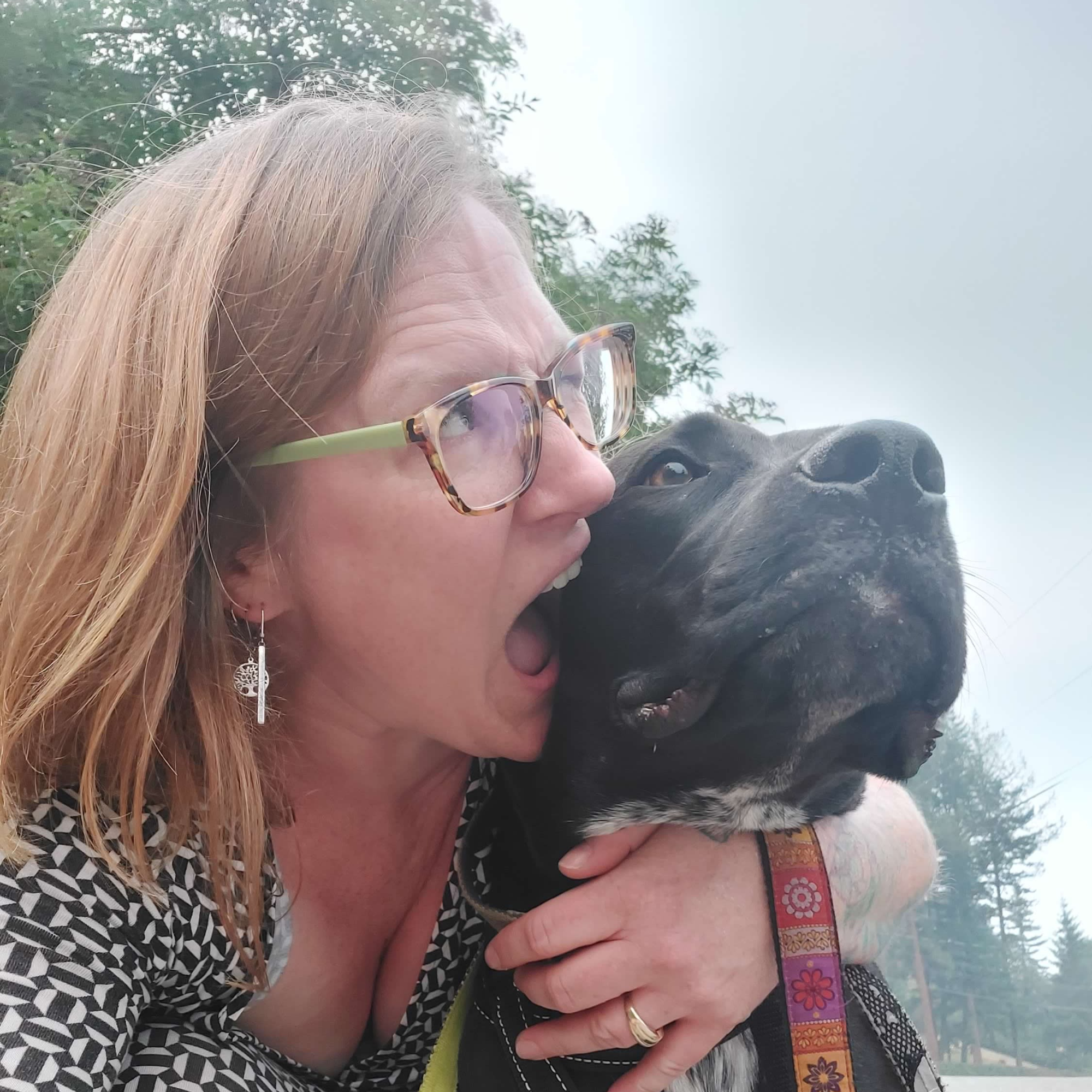 : A blonde woman wearing glasses pretends to bite her black dog in a selfie-style picture
