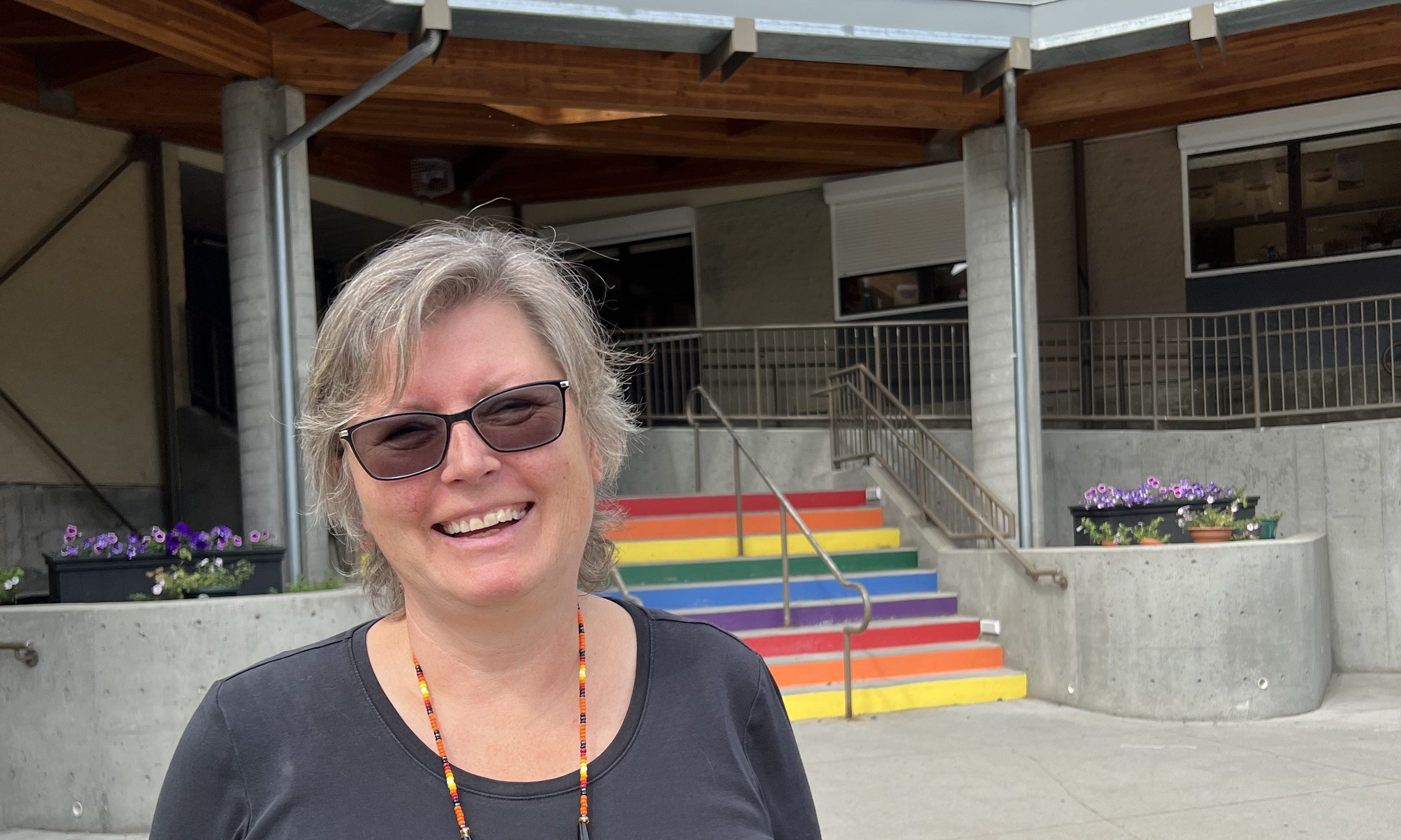 A woman with short grey hair and sunglasses in a blue shirt and red necklace stands in front of a school with rainbow steps.