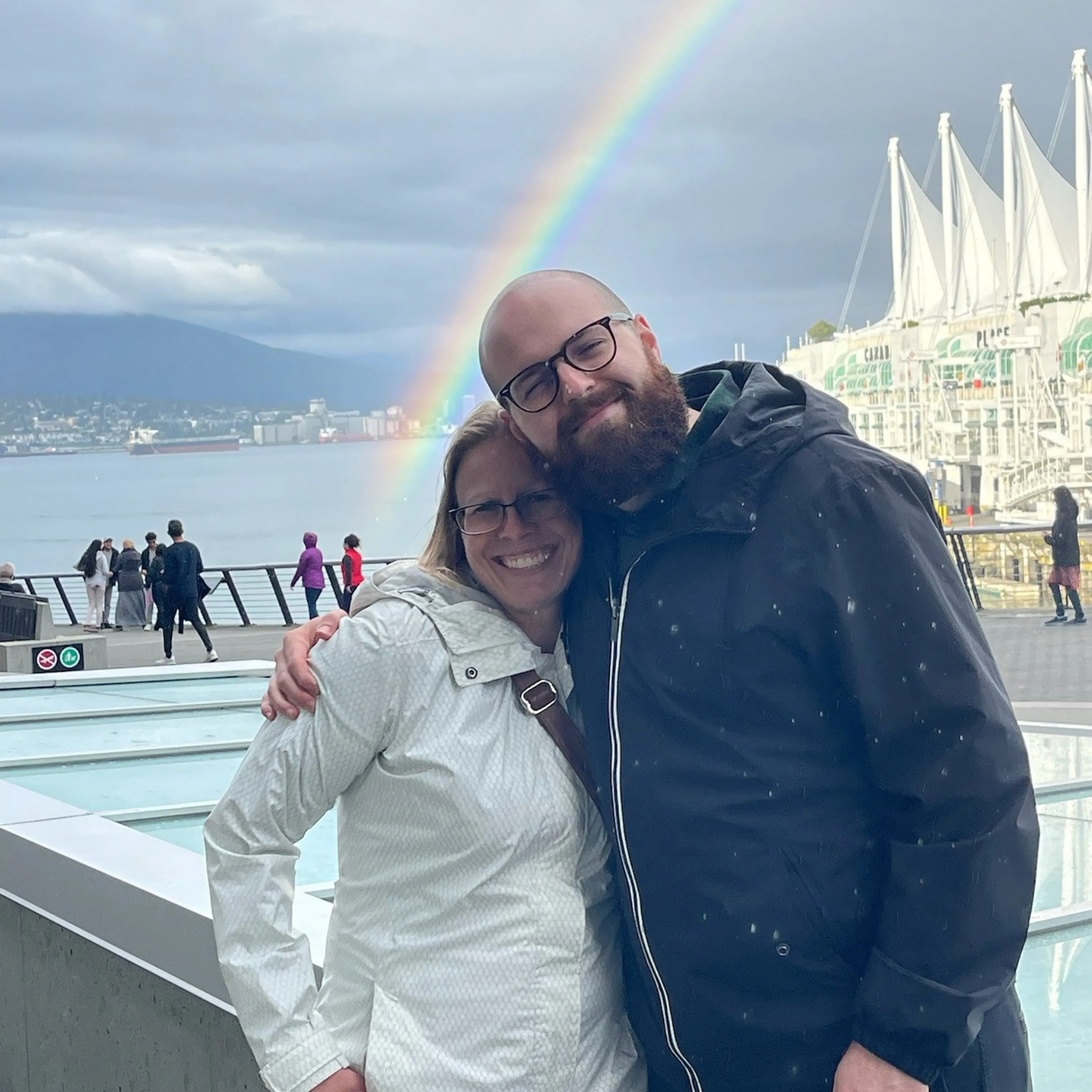Two people embrace for a picture with big smiles while standing in front of a fountain, a beach and a rainbow.