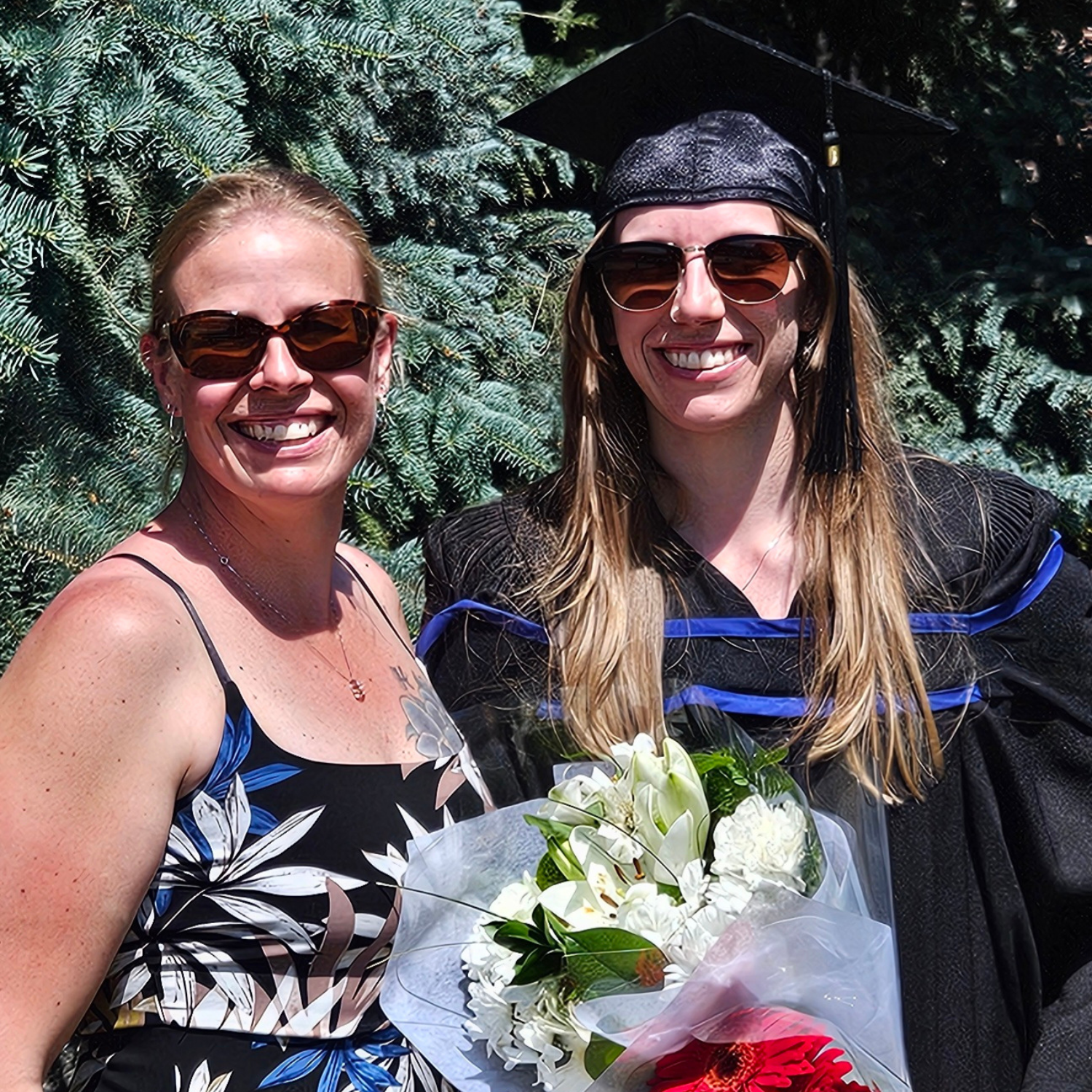 Two people stand side by side with big smiles. The person on the right is in a graduation cap and gown and they are holding flowers. 