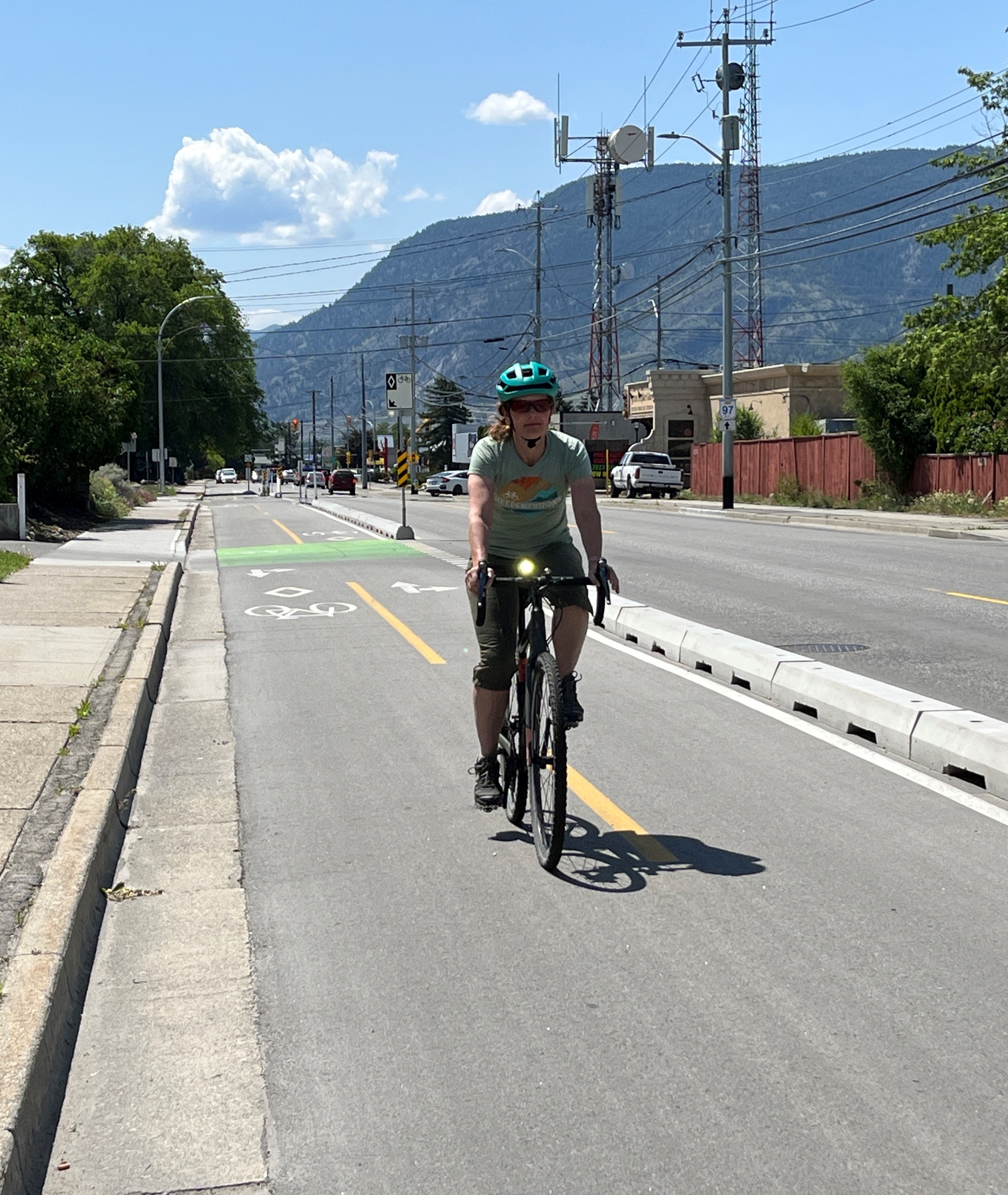 A person in a green bike helmet, green tshirt and short pants rides in a bike lane with mountains in the background