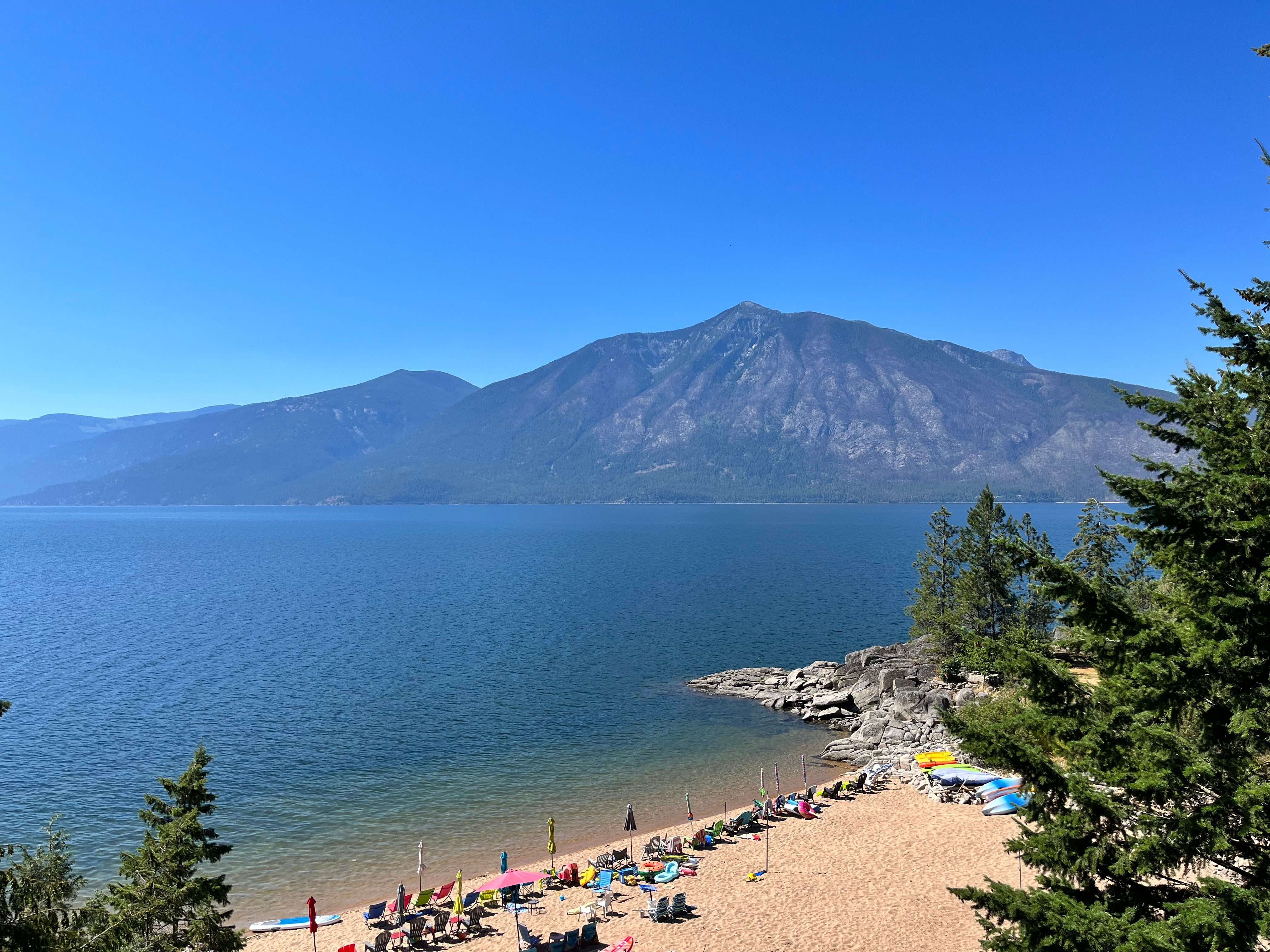 An overhead view of a blue lake and white sand beach with pine trees in the foreground and a mountain in the background