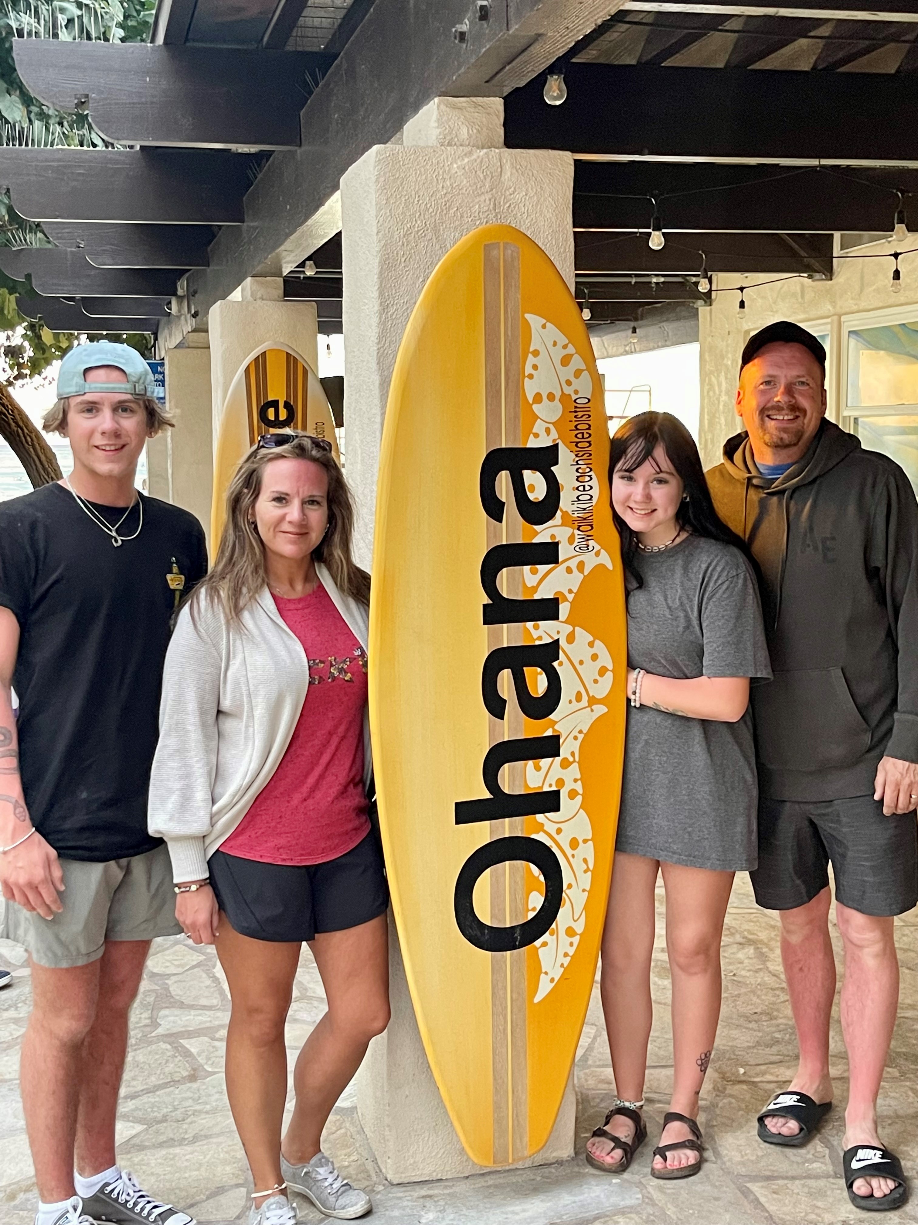 Two adults and two younger people pose for picture while standing underneath an awning and holding a large surfboard that reads Ohana.