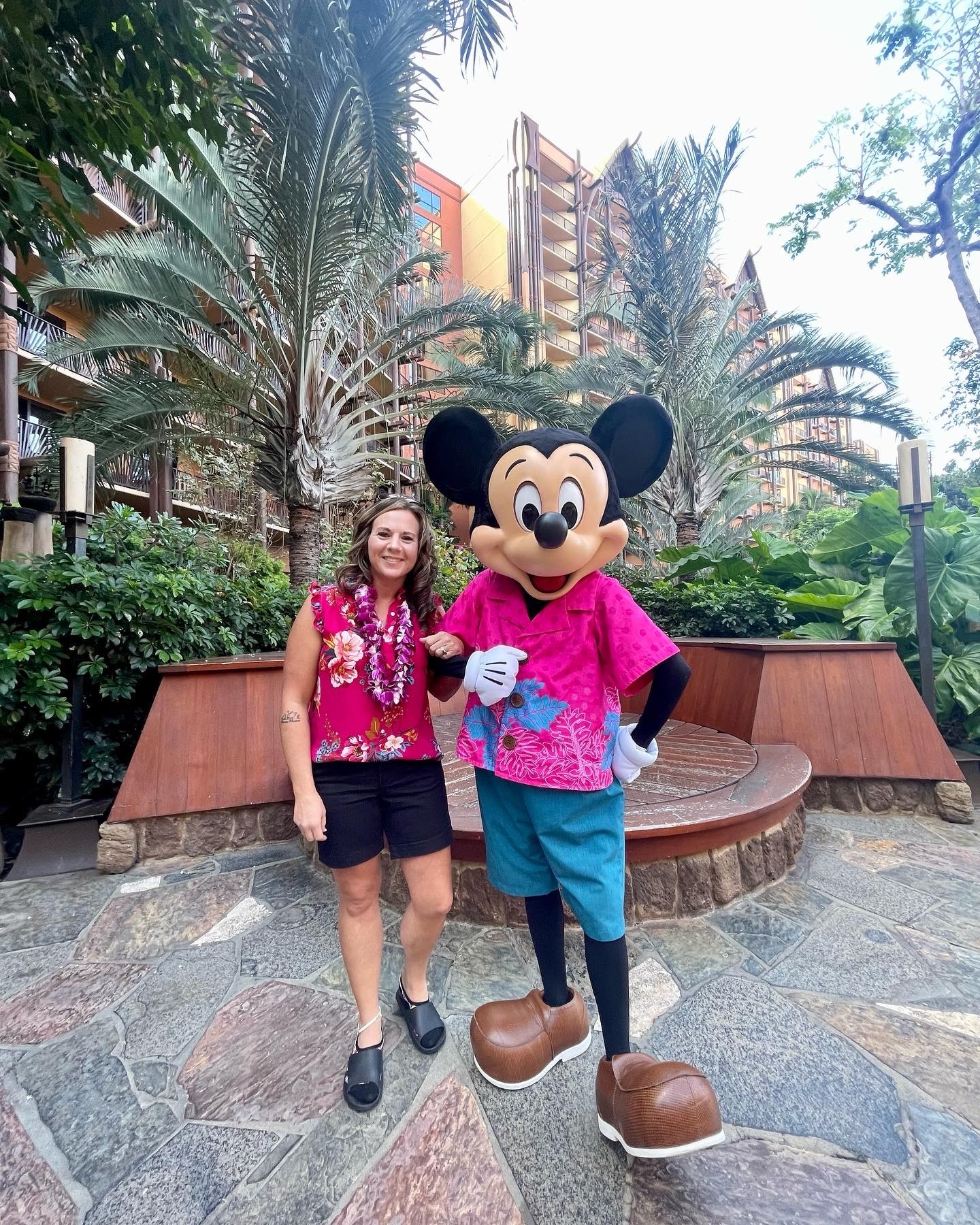 : A woman in Hawaiian attire poses for a picture outside of a resort while linking hands with someone in a Mickey Mouse costume that includes a Hawaiian shirt.