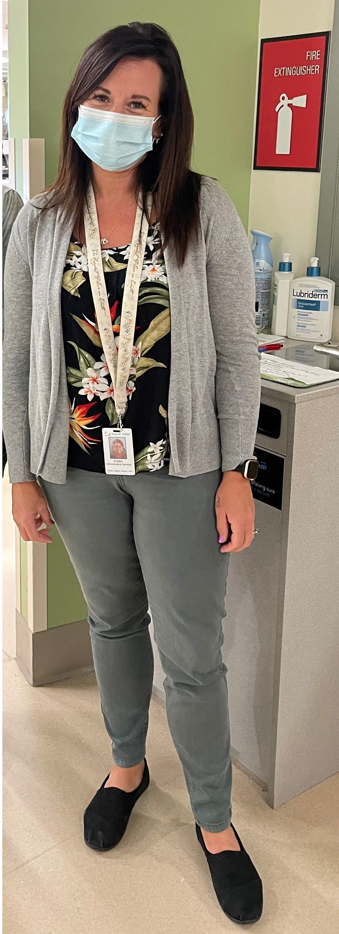 A smiling woman dressed in casual office attire, wearing a mask and a name tag, poses for a picture while standing up.