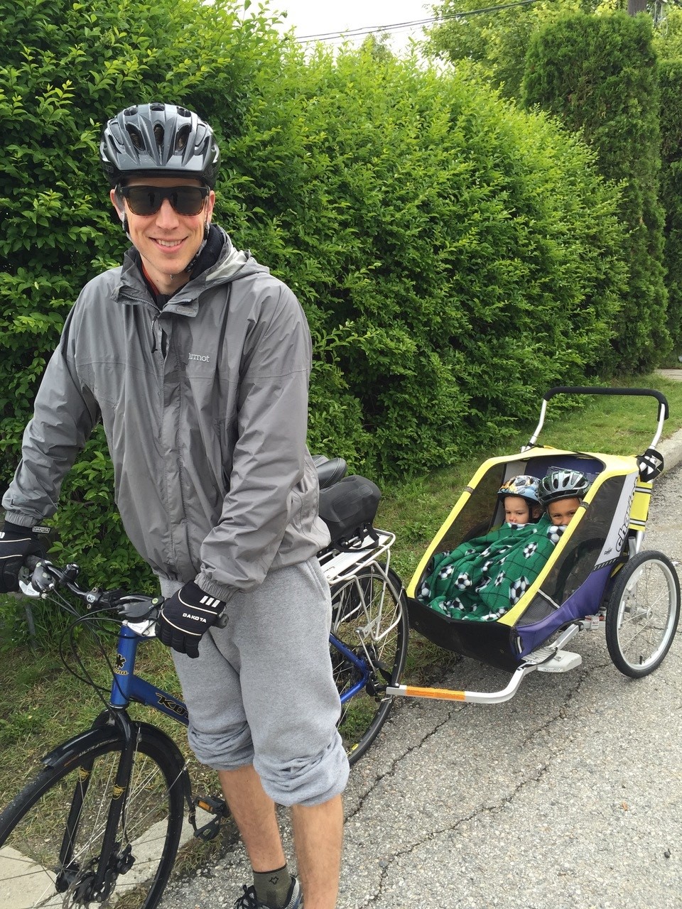 A man in a grey jacket, track pants and cycling helmet leans on a blue bike attached to a yellow trailer in which two children covered in a green blanket sit