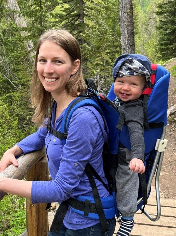 A person wearing a long-sleeved blue top and carrying a smiling baby in a backpack stands on a bridge in a forest.