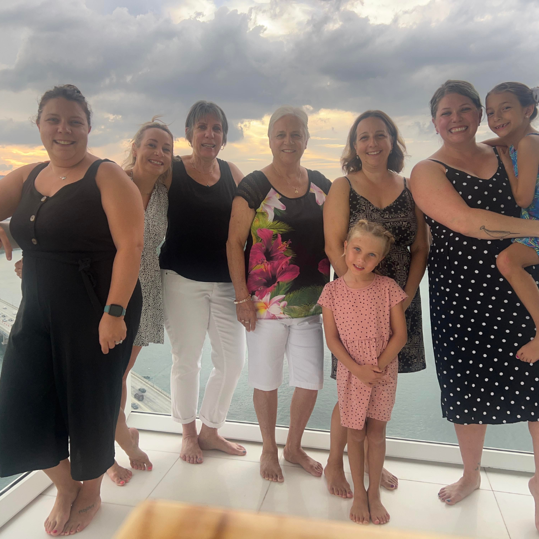 a group of Caucasian women and two girls ranging in age smile together while visiting a waterfront location.