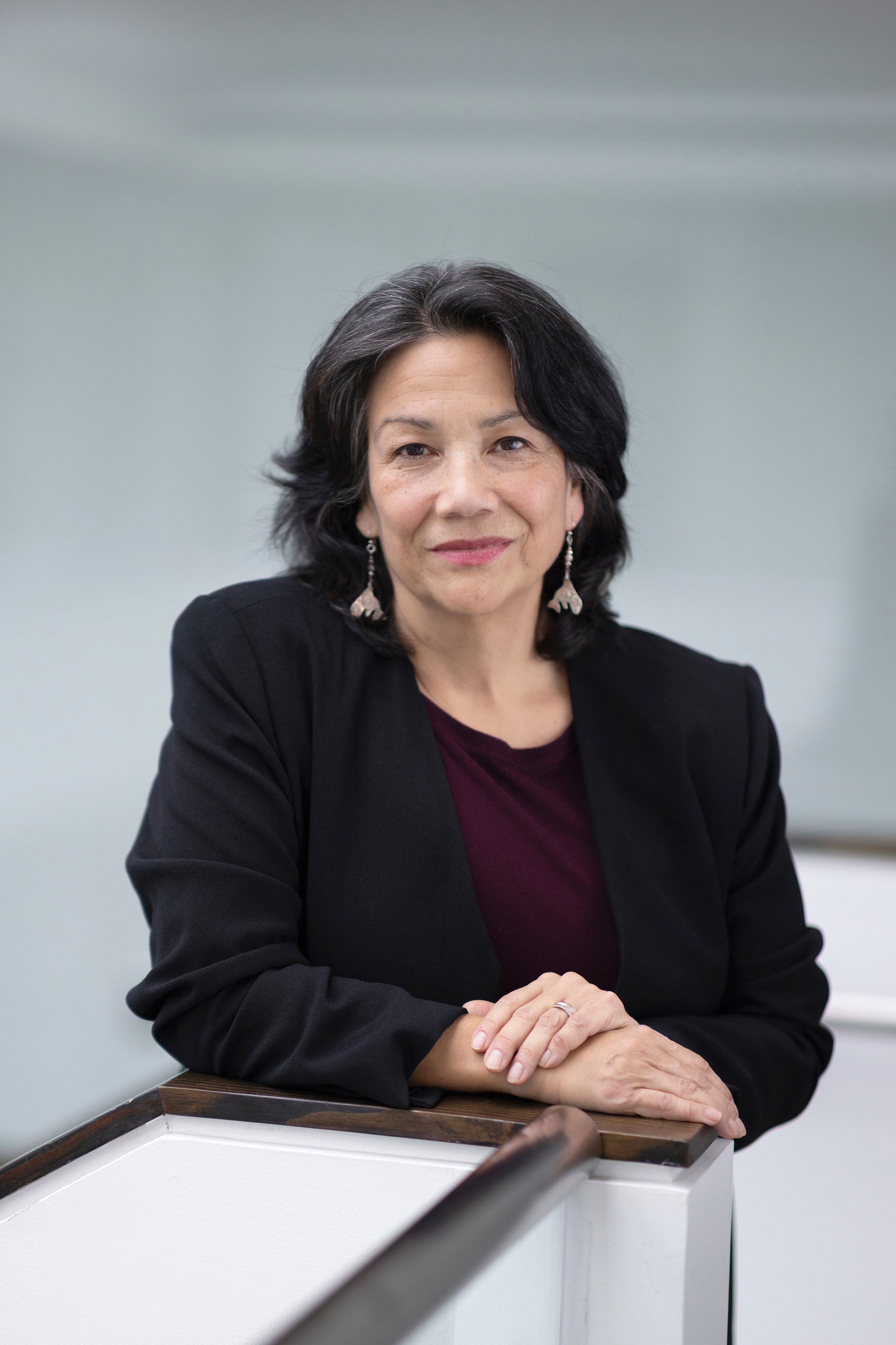 Profile shot of a person with mid length black hair wearing earrings, a dark red shirt and black blazer.