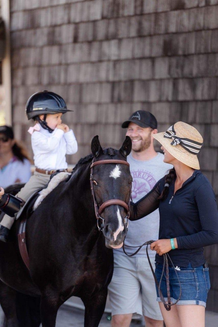 A young child wearing a helmet sits on a black horse with a white diamond on its forehead next to two adults, both in hats